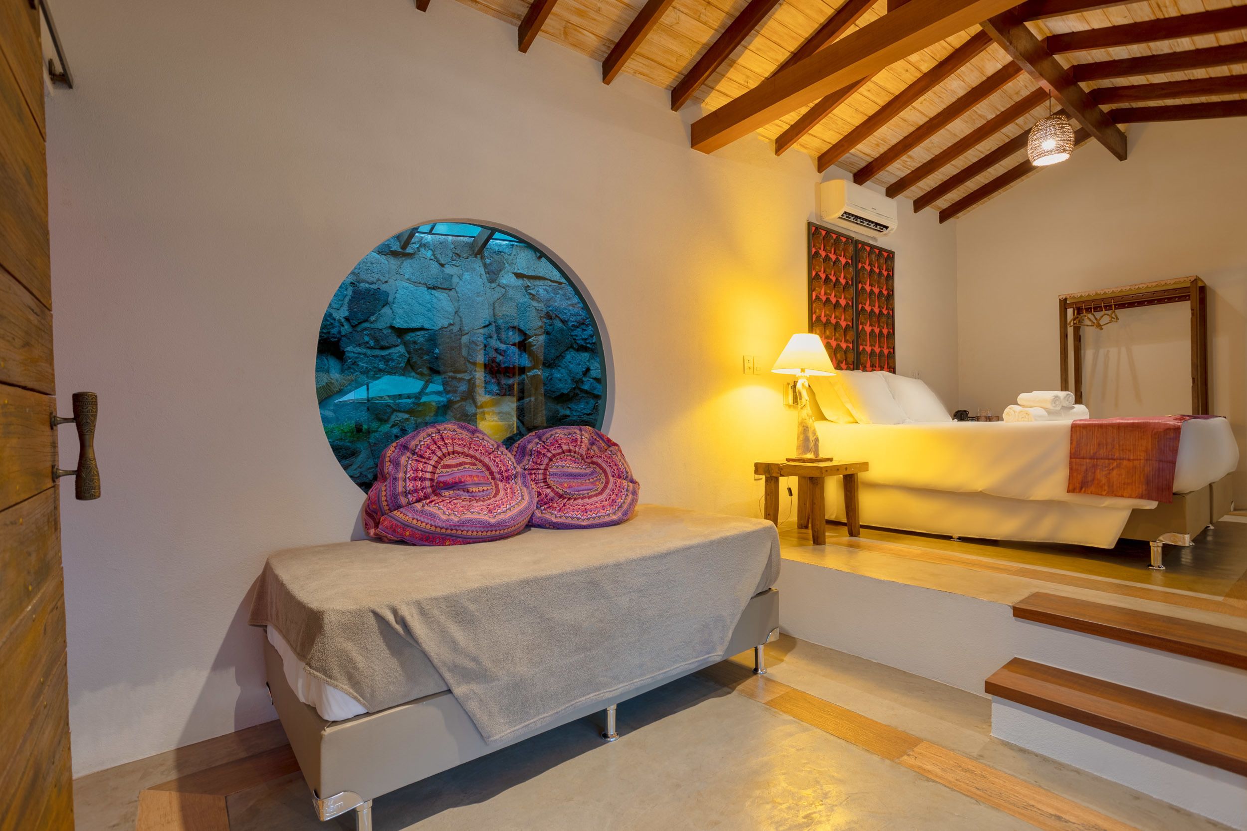 Una camera da letto con un muro di pietra circolare, un letto con coperta bianca, un comodino con una lampada e una scala che conduce a un'altra stanza.