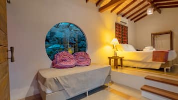 Una camera da letto con un muro di pietra circolare, un letto con coperta bianca, un comodino con una lampada e una scala che conduce a un'altra stanza.