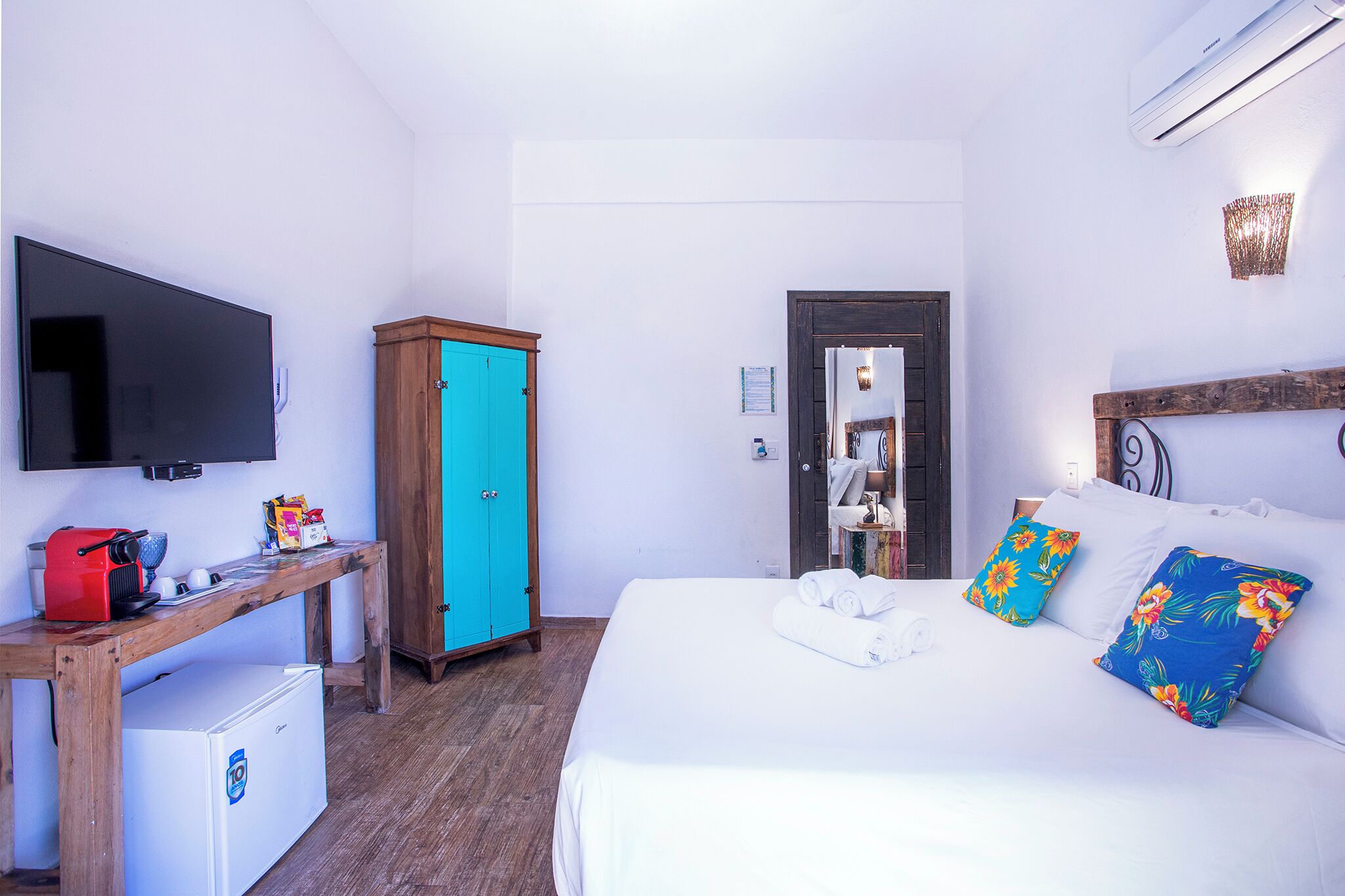 A hotel room with a bed, a TV, a desk with a coffee maker, a wardrobe, and a mirror.