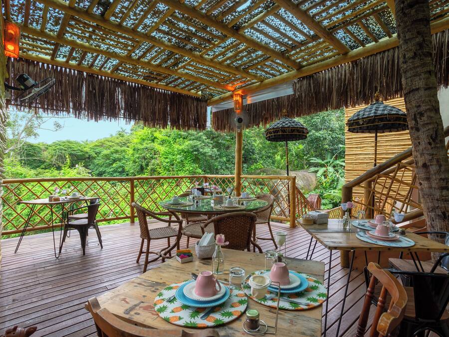 A wooden deck with tables set for tea service, surrounded by wicker chairs, overlooking lush greenery.