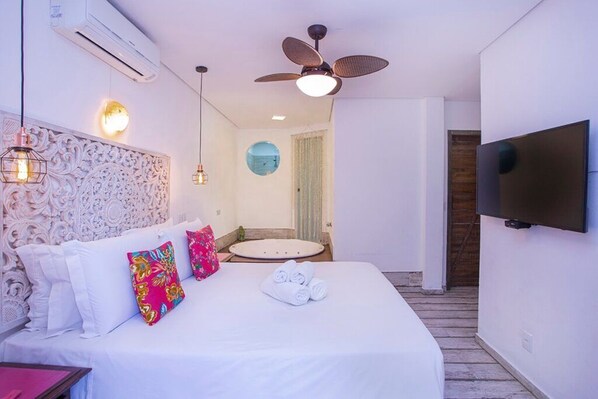 A hotel room with a bed, a ceiling fan, a TV, and a bathroom with a round mirror.