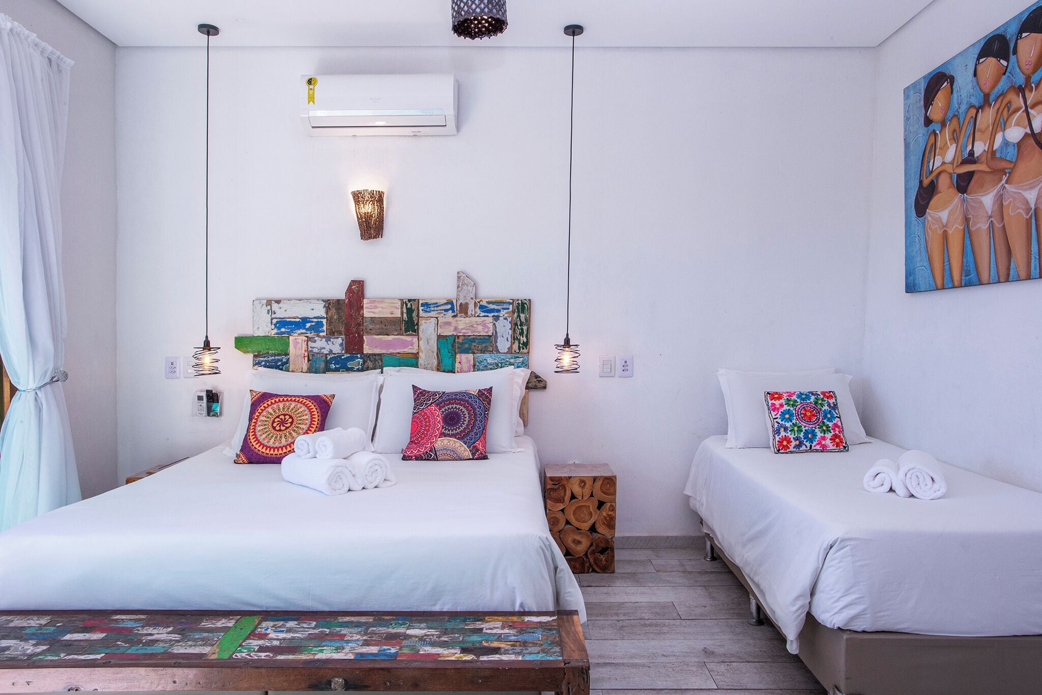 Habitación de hotel con una cama grande, dos camas más pequeñas, un cabecero de madera, una obra de arte colorida y una mesa de madera.