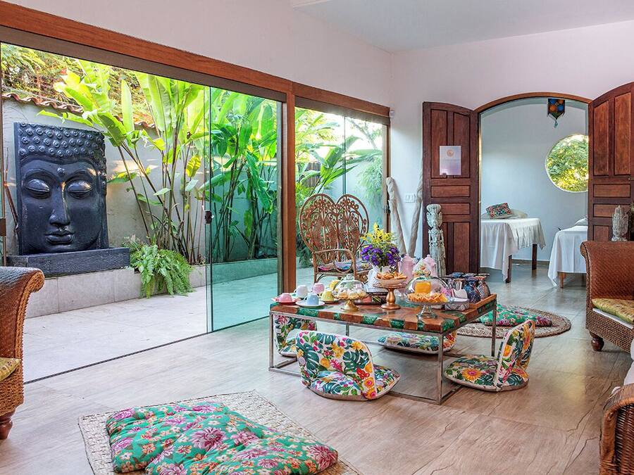 A room with a glass door leading to a garden, wicker furniture, a table with floral cushions, and a Buddha statue.