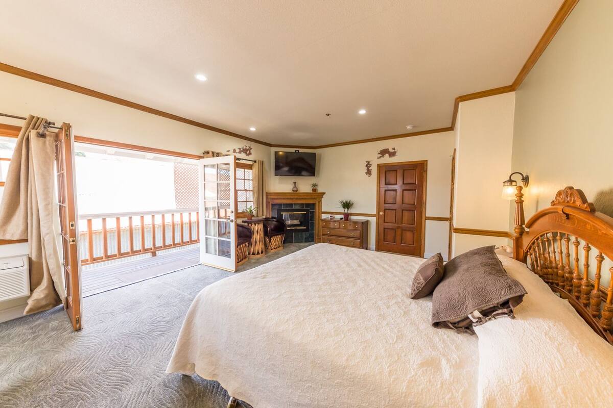 A bedroom with a large bed, a wooden headboard, a TV mounted on the wall, a fireplace, and a balcony with curtains.