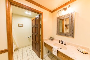 A bathroom with a wooden door, a white sink with two faucets, a mirror with an ornate frame, and a small plant on the counter.