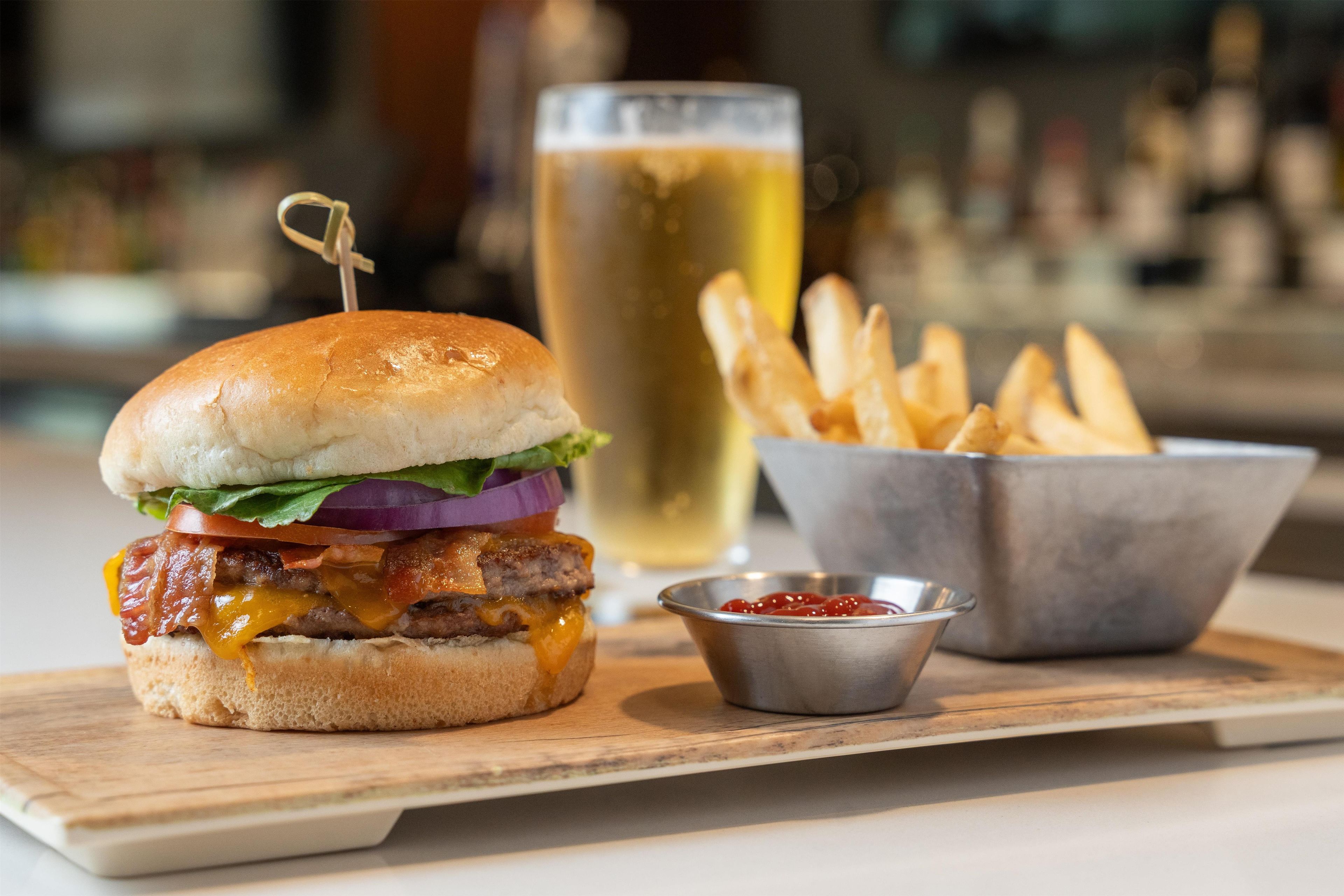 Una hamburguesa con lechuga, tomate y cebolla morada, un vaso de cerveza y un bol de papas fritas sobre una tabla de madera.