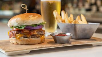 Una hamburguesa con lechuga, tomate y cebolla morada, un vaso de cerveza y un bol de papas fritas sobre una tabla de madera.