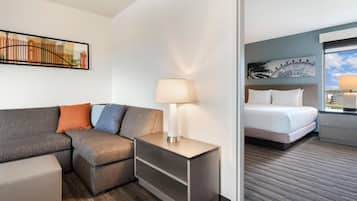 A hotel room with a sofa, a bedside table with a lamp, a bed, and a painting on the wall.