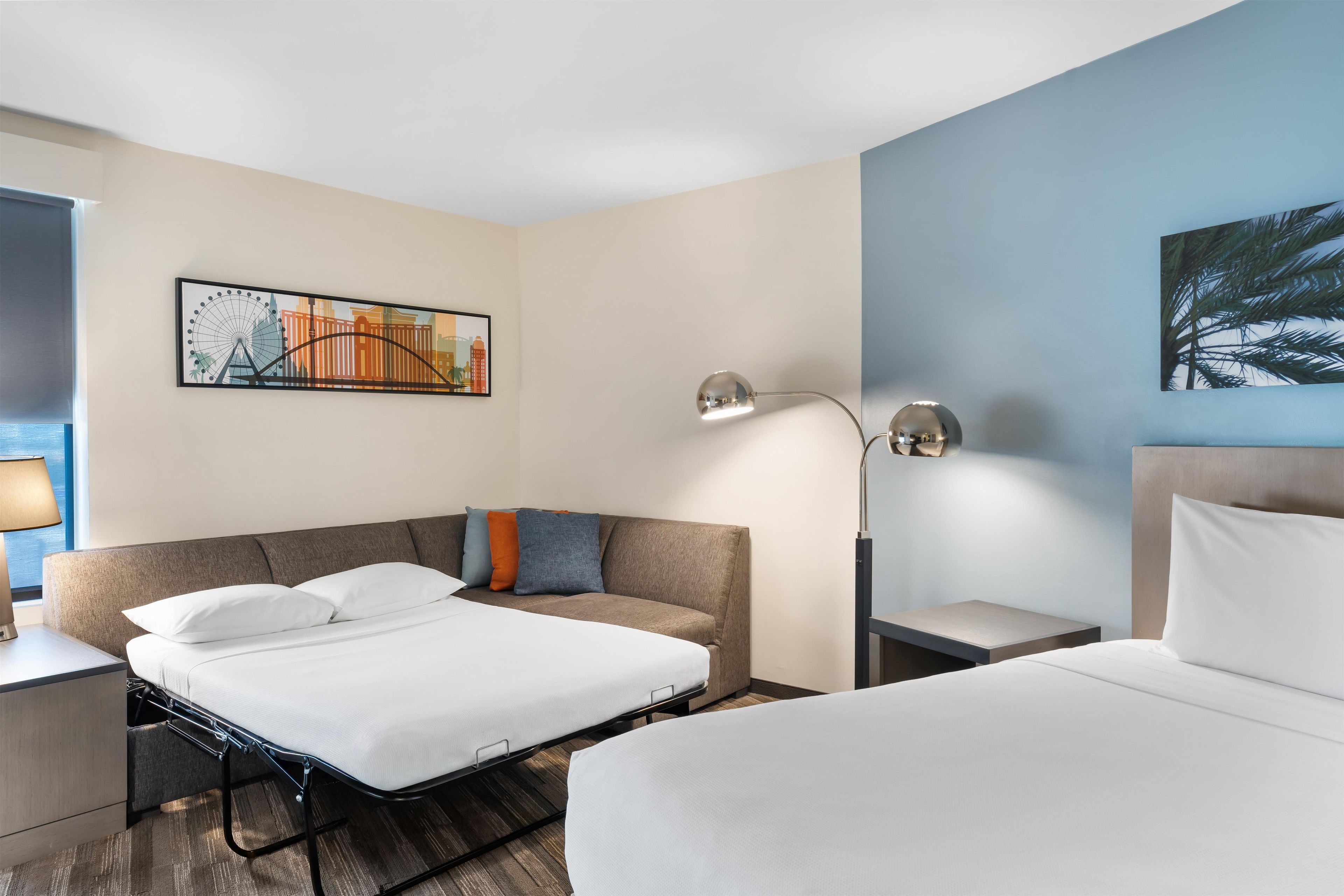 A hotel room with a bed, a sofa, a nightstand, a lamp, and a wall art piece.