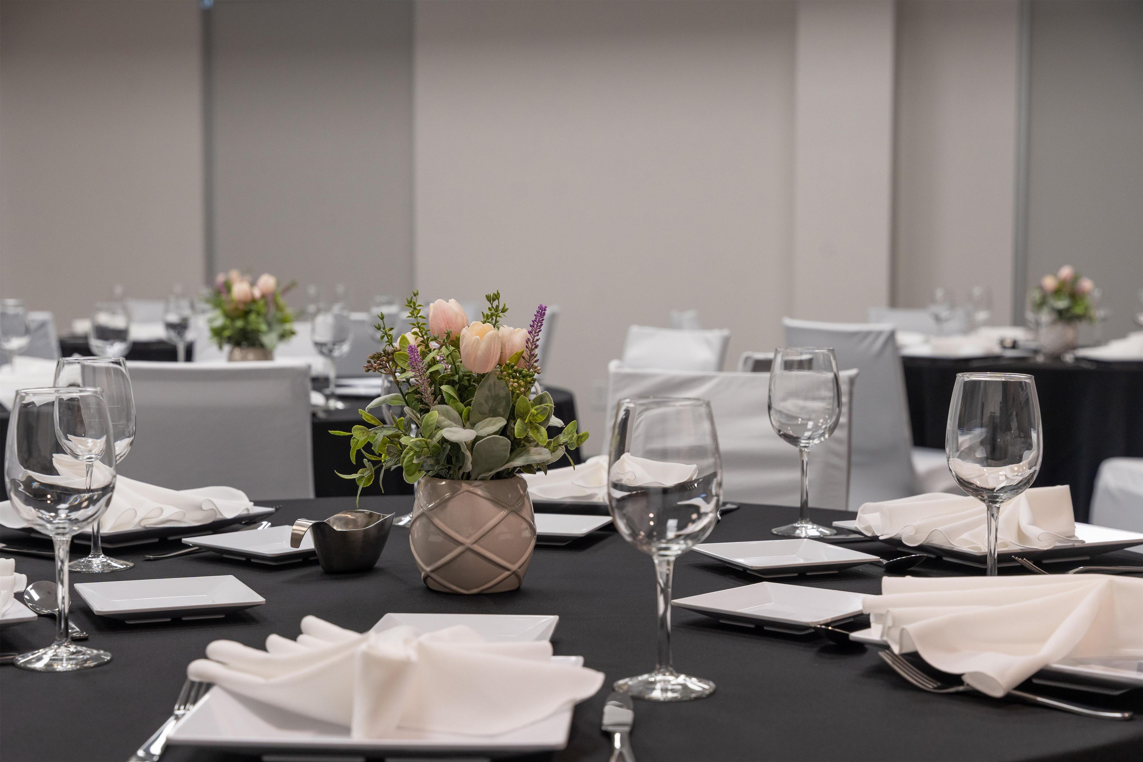 Una sala de conferències amb taules preparades per a una reunió, cadascuna amb un got de cristall transparent, una tovallola blanca plegada i un vas petit amb flors.