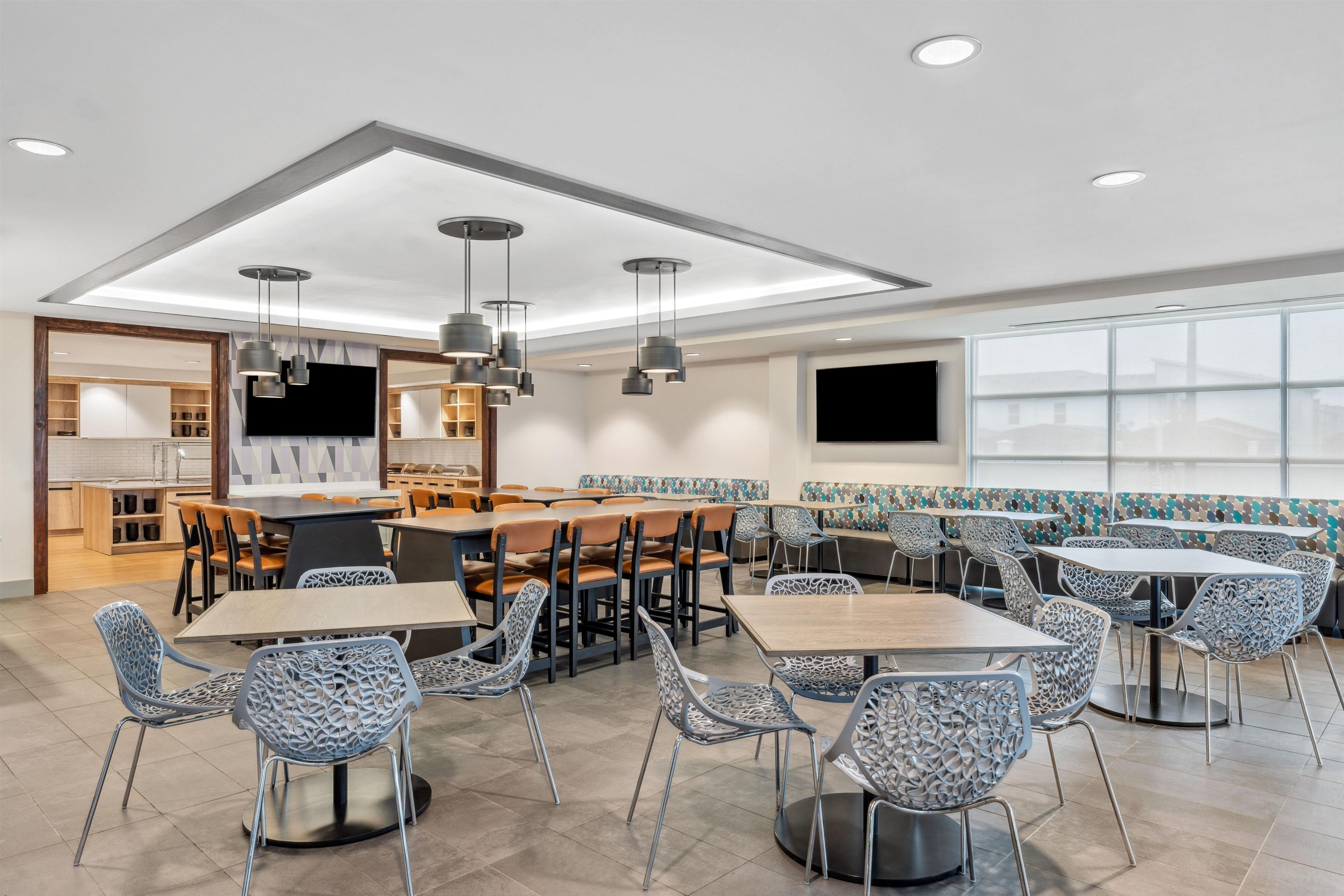 A modern dining area with multiple tables and chairs, a kitchen in the background, and large windows.
