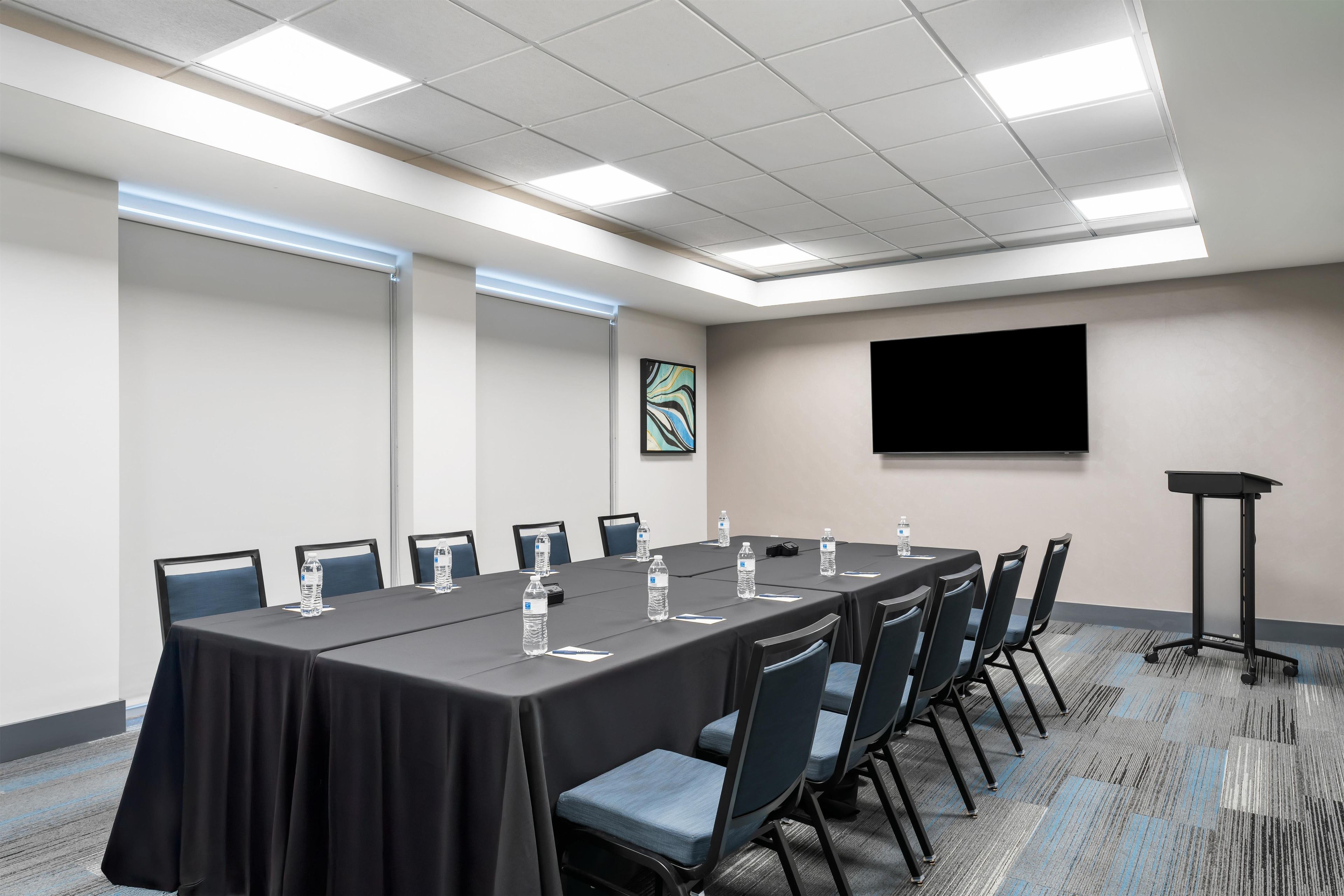 Un salón de conferencias con una larga mesa, sillas, botellas de agua y una pantalla de proyector.