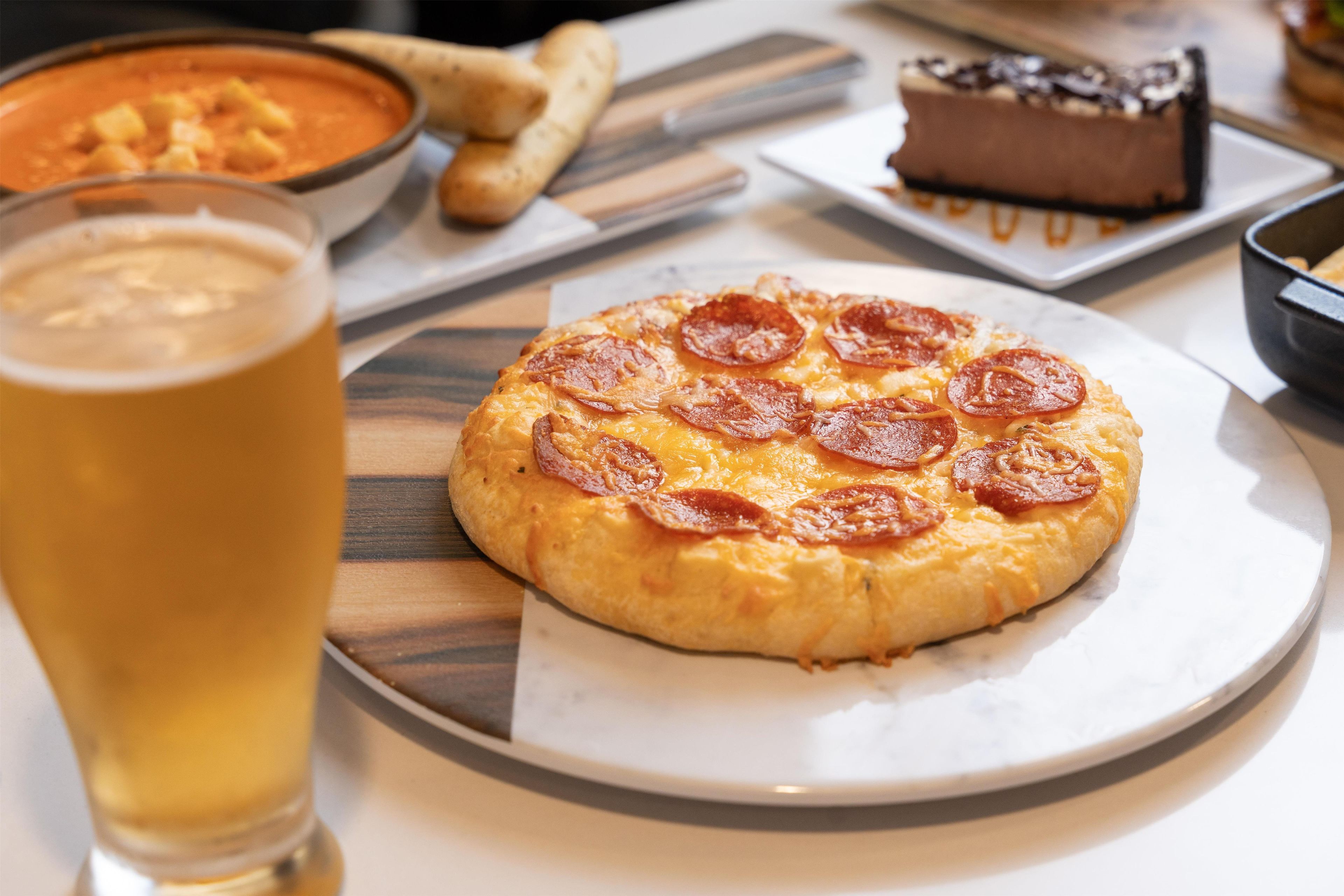 A pizza with pepperoni on a plate, a glass of beer, and a dessert on a table.