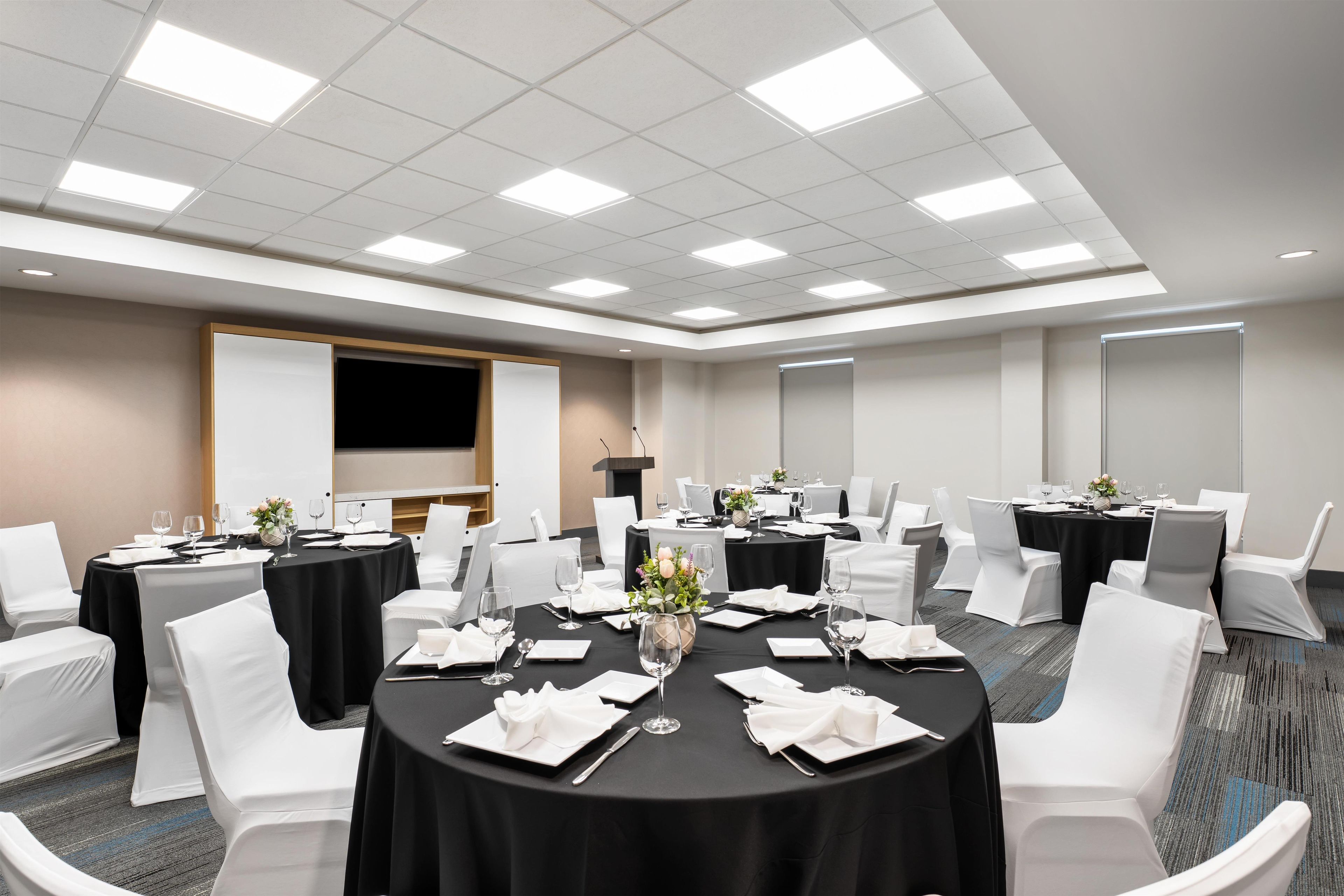 Un salón de conferencias con mesas redondas, sillas blancas y una pantalla de presentación.