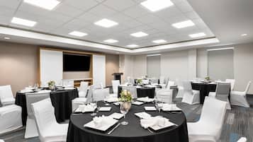 Un salón de conferencias con mesas redondas, sillas blancas y una pantalla de presentación.