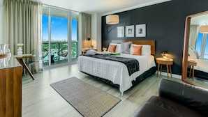 A modern hotel room with a large bed, a sitting area, a desk, and a balcony with a city view. - iCoconutGrove Vacation Rentals (Miami)
