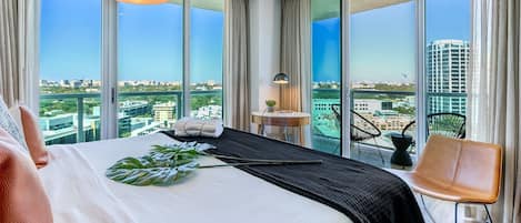 A modern hotel room with a large bed, a view of the city, and a balcony with a chair.