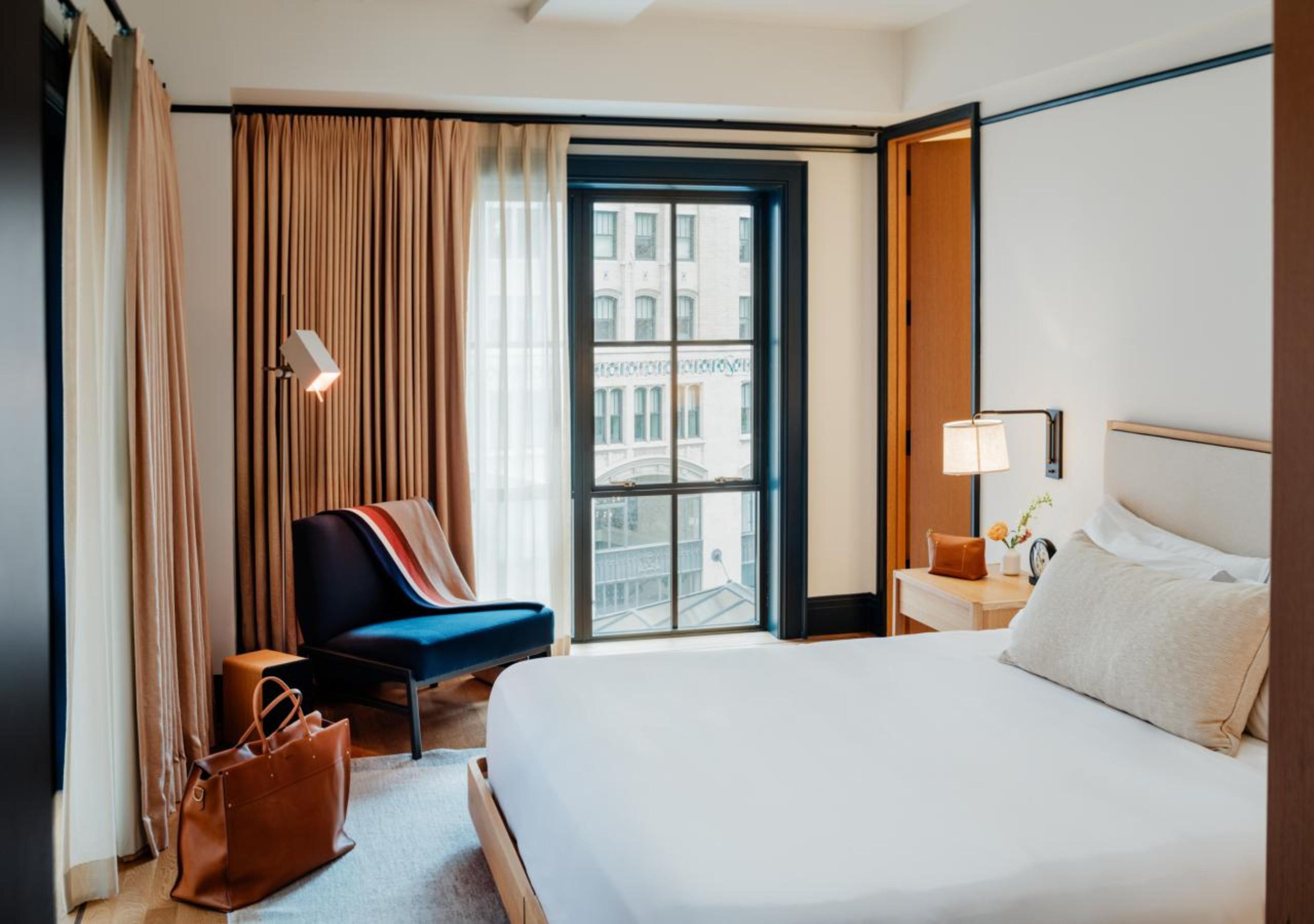 A hotel room with a bed, a chair, a nightstand, a lamp, and a view of a building through a window.