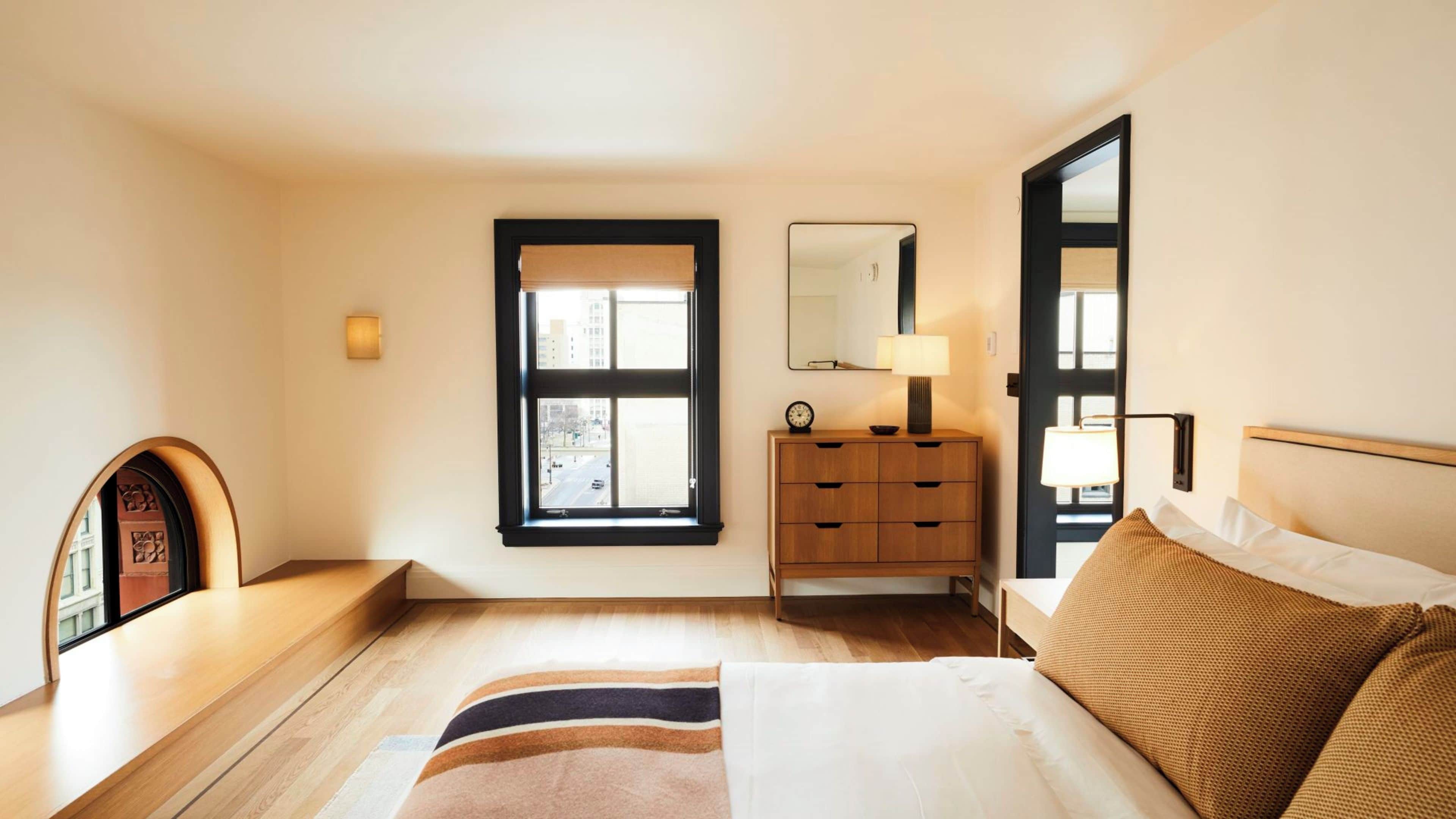 Habitación de hotel con cama, cómoda de madera, mesita de noche, espejo y ventana con vista a la ciudad.