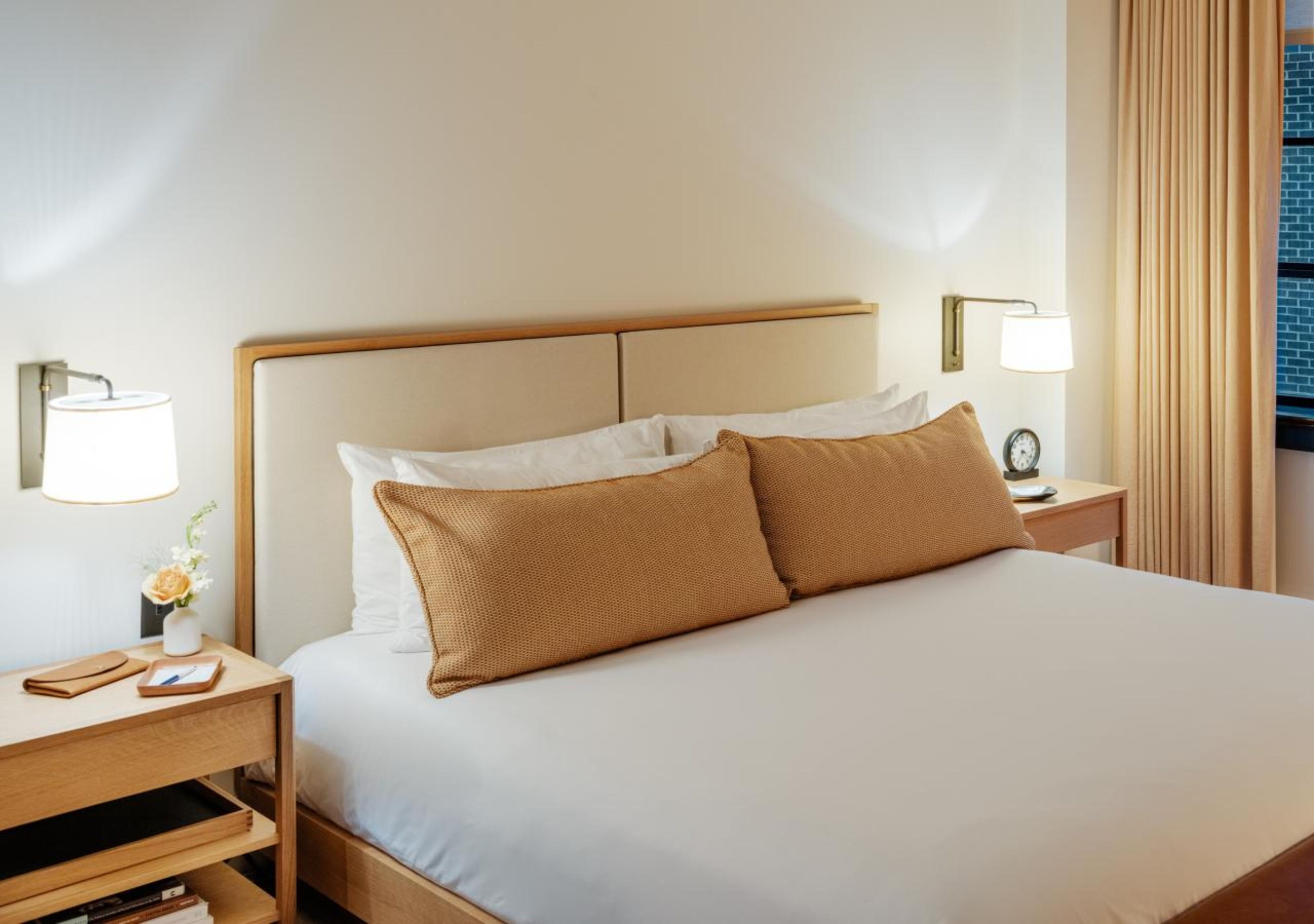 A hotel room with a bed, two bedside tables, a lamp, a clock, and a window with curtains.