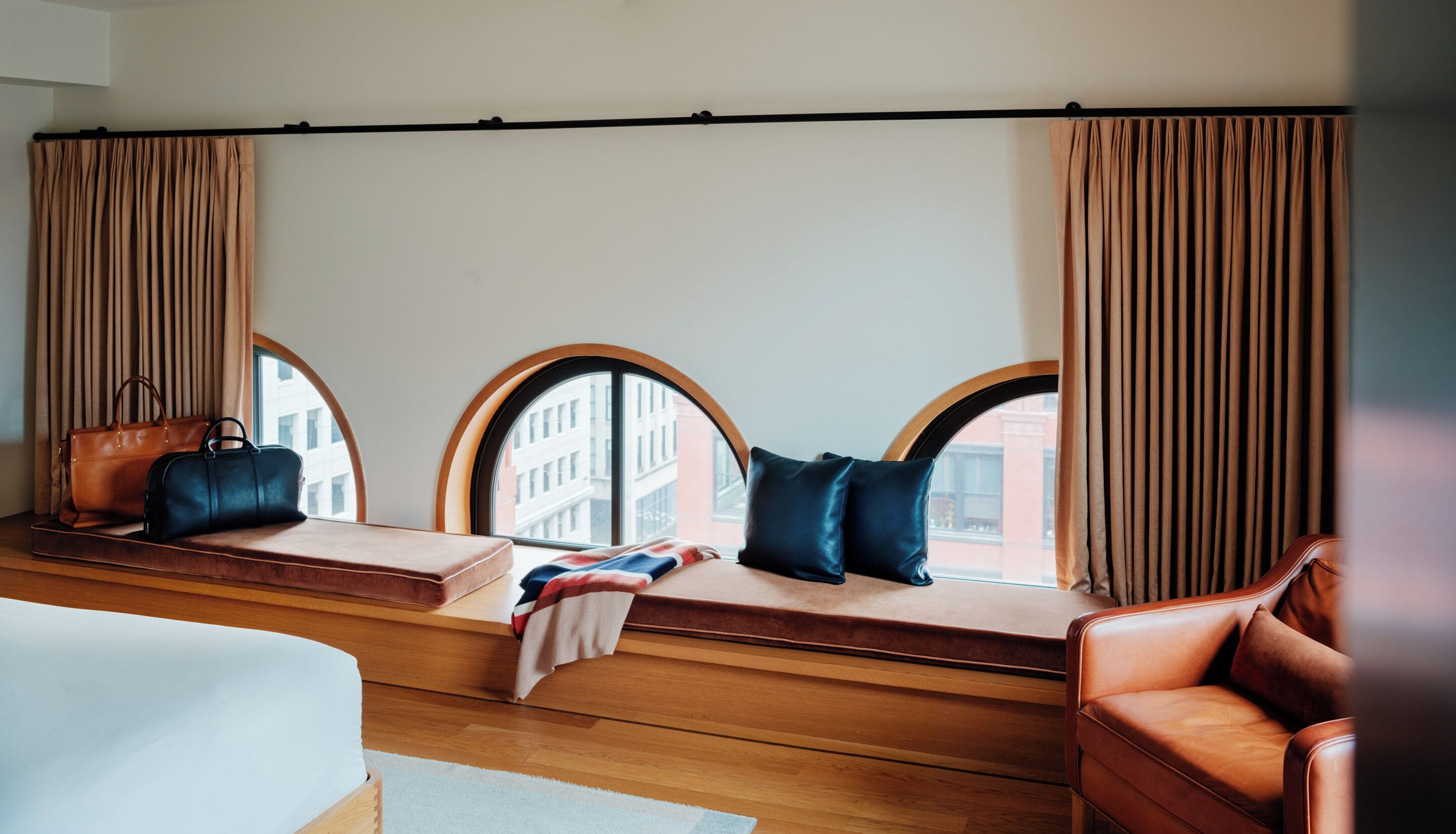 A hotel room with a large window seat, leather armchairs, and a view of buildings.