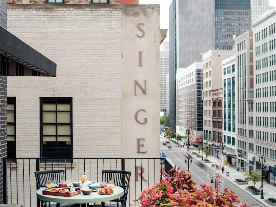 Una mesa con alimentos para el desayuno en un balcón con vistas a una calle y un edificio cuya fachada muestra 'SINGER'.