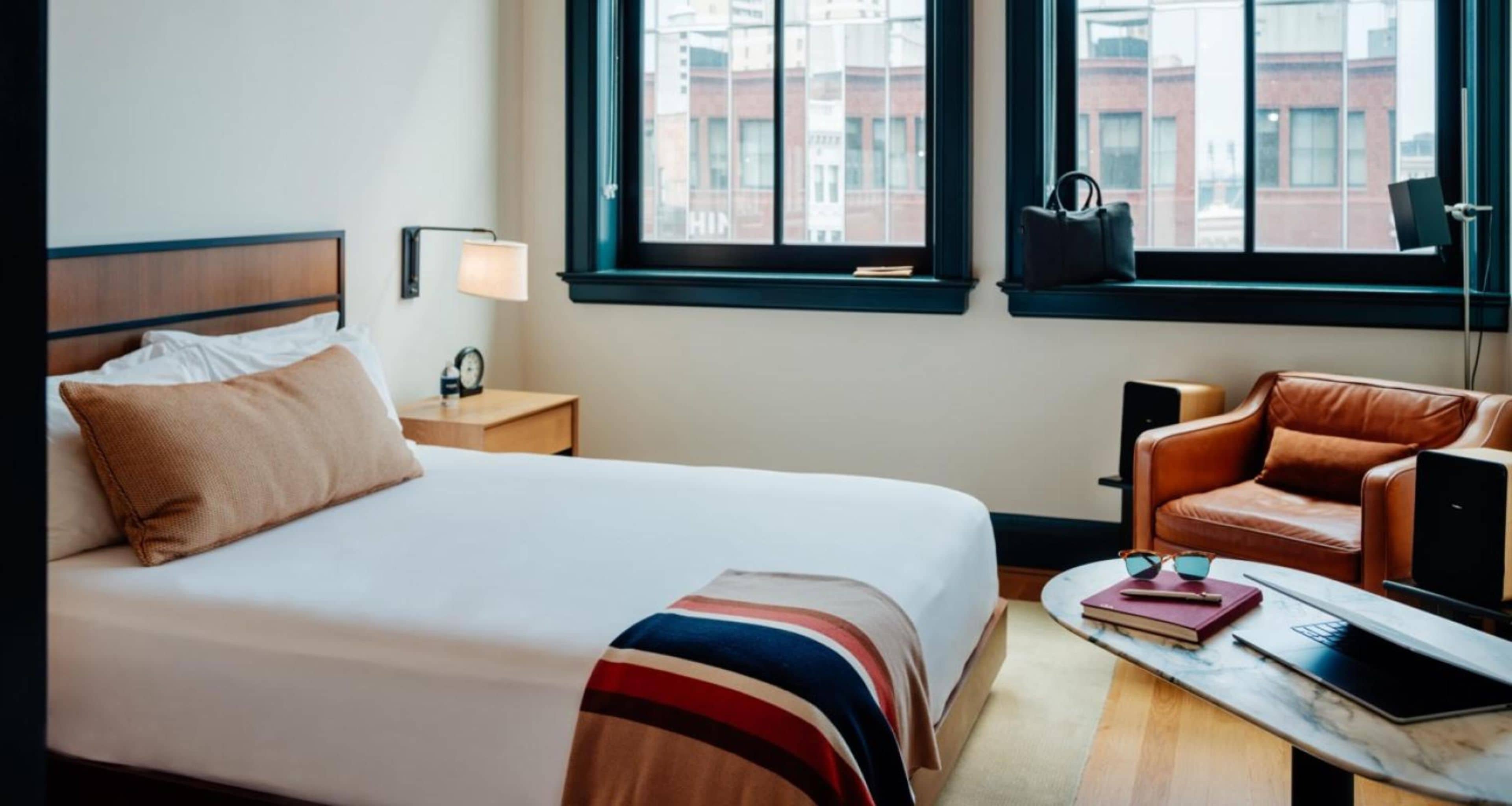 A hotel room with a bed, a nightstand, a lamp, a chair, a desk, and a window with a view of buildings.