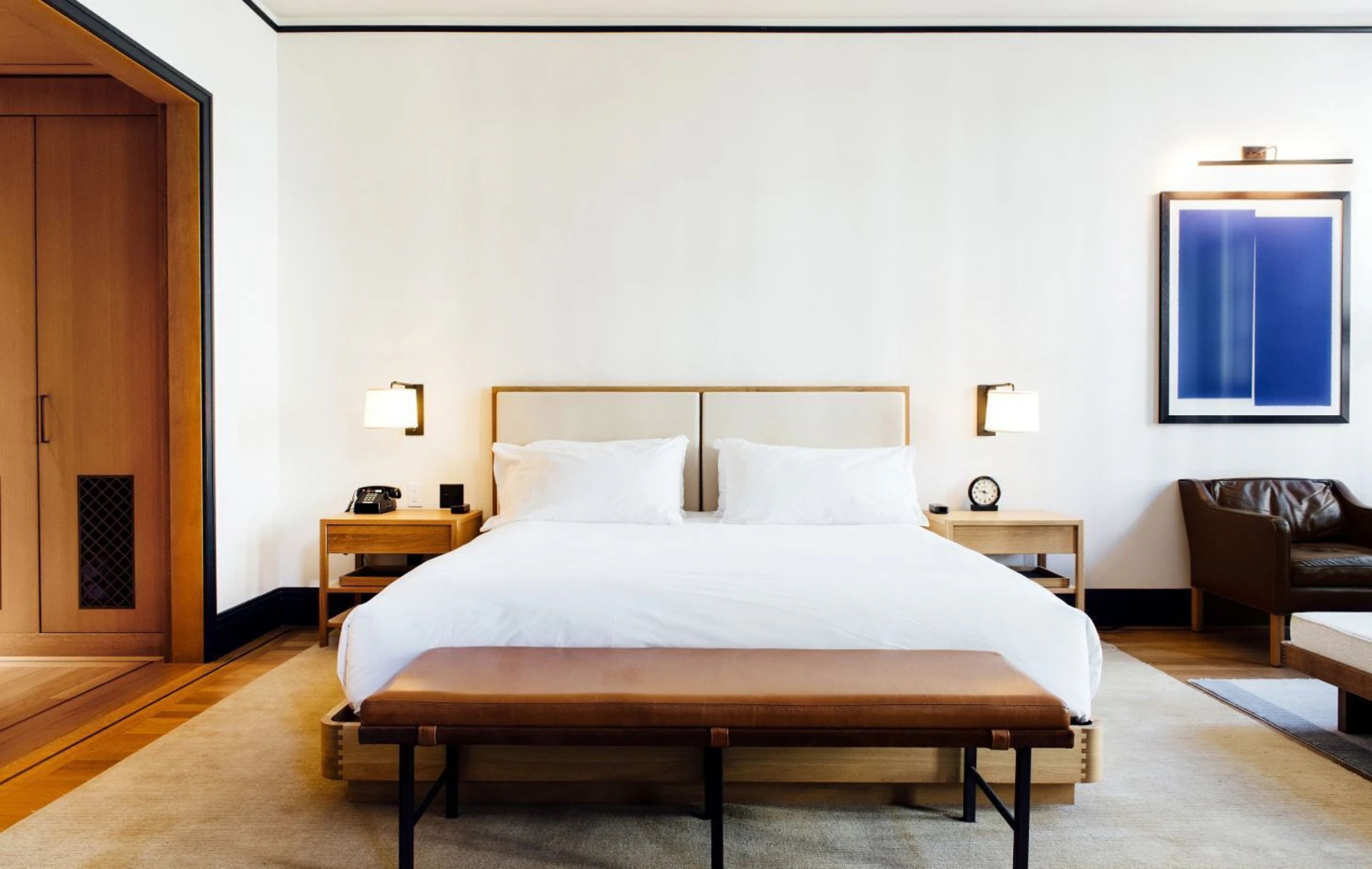 Habitación de hotel con una cama grande, mesitas de noche, un banco, una silla y una obra de arte colgada en la pared.