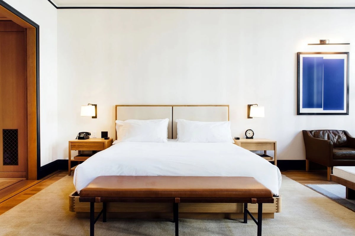 A neatly arranged hotel room with a large bed, bedside tables, a bench, a wooden wardrobe, and a framed artwork on the wall.