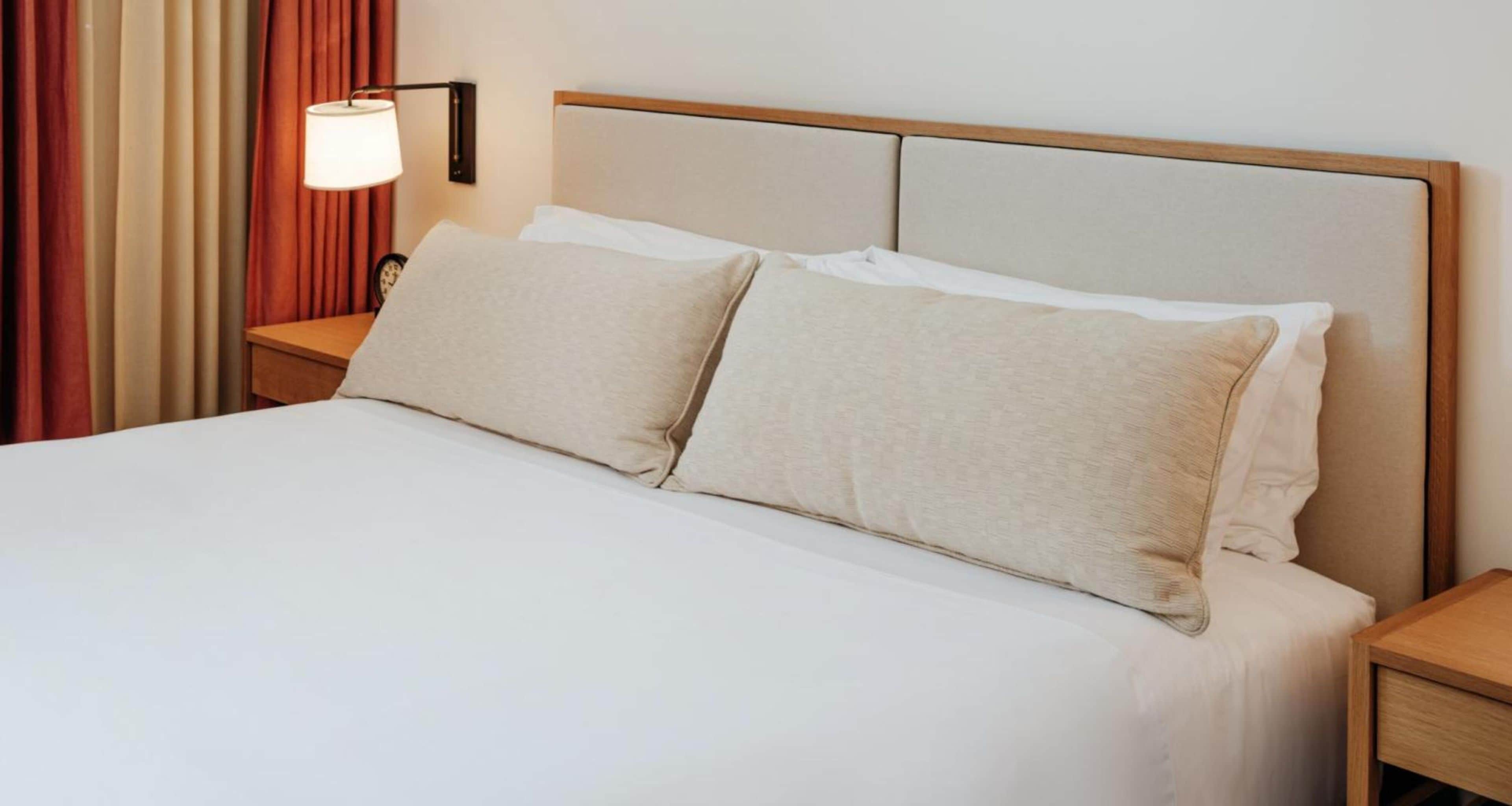 Habitació d'hotel amb llit, dos coixins, una taula de nit, una làmpada i cortines.
