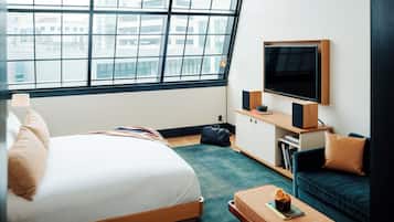 A modern hotel room with a large window, a bed, a TV, a sofa, and a small table with a book and a vase.