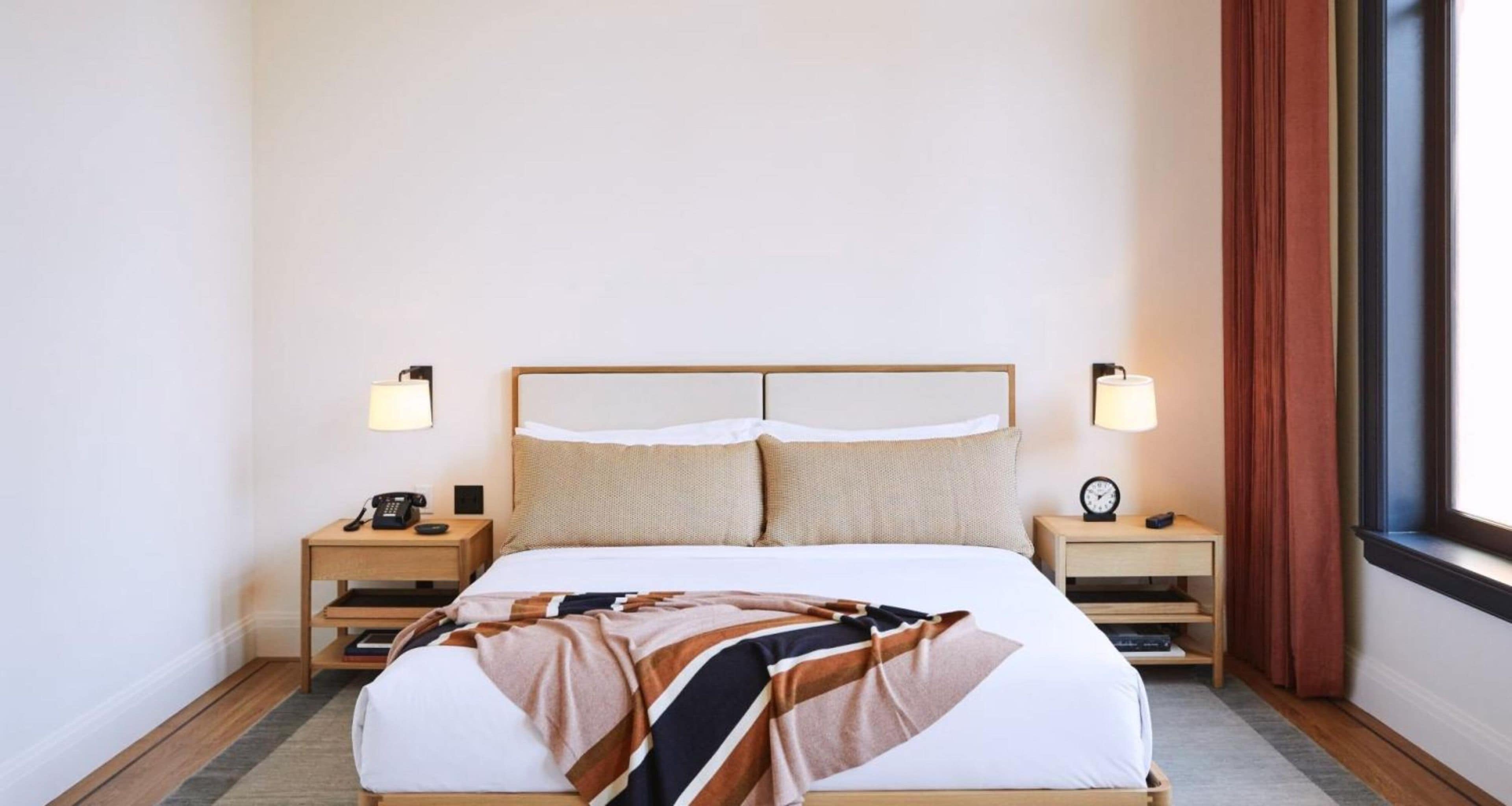 A hotel room with a bed, bedside tables, lamps, a clock, and a window with curtains.