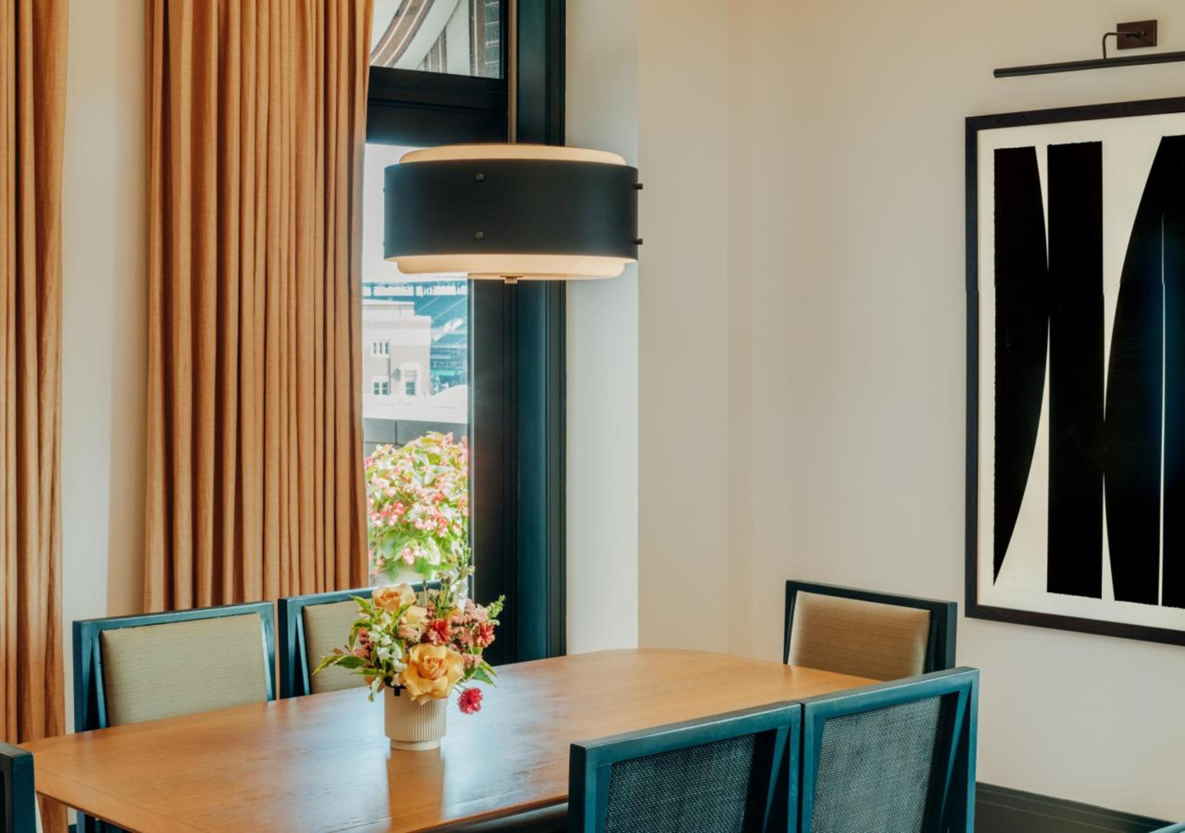 Un comedor con una mesa de madera, sillas, un jarrón de flores, una obra de arte grande con marco negro y una ventana con cortinas.