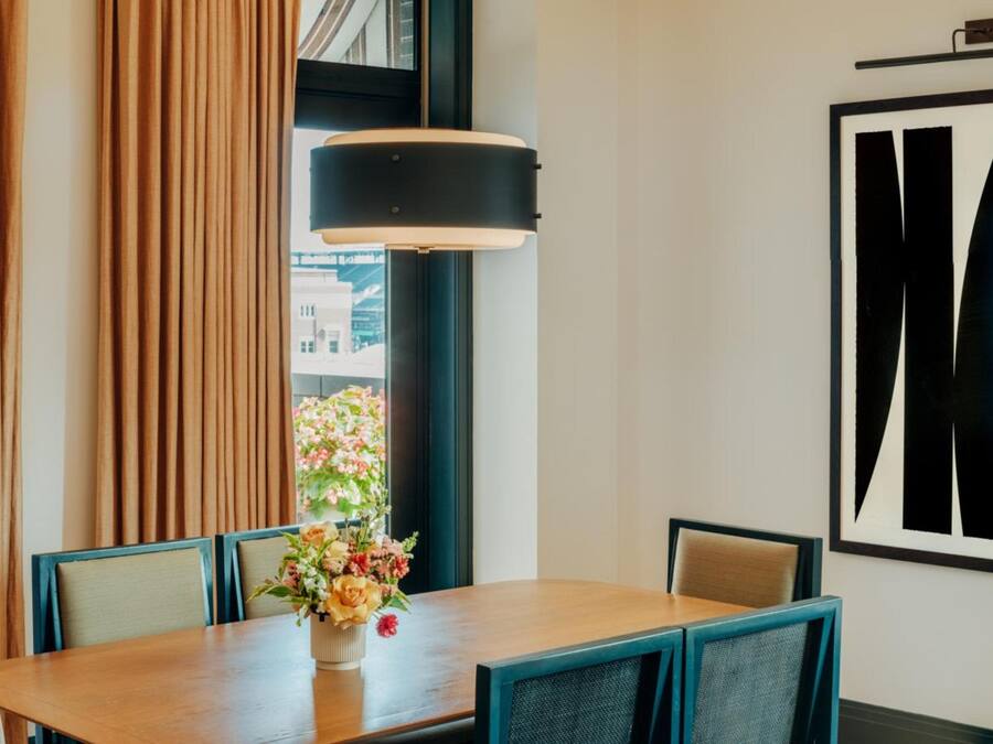 Un comedor con una mesa de madera, sillas, un jarrón de flores, una obra de arte grande con marco negro y una ventana con cortinas.