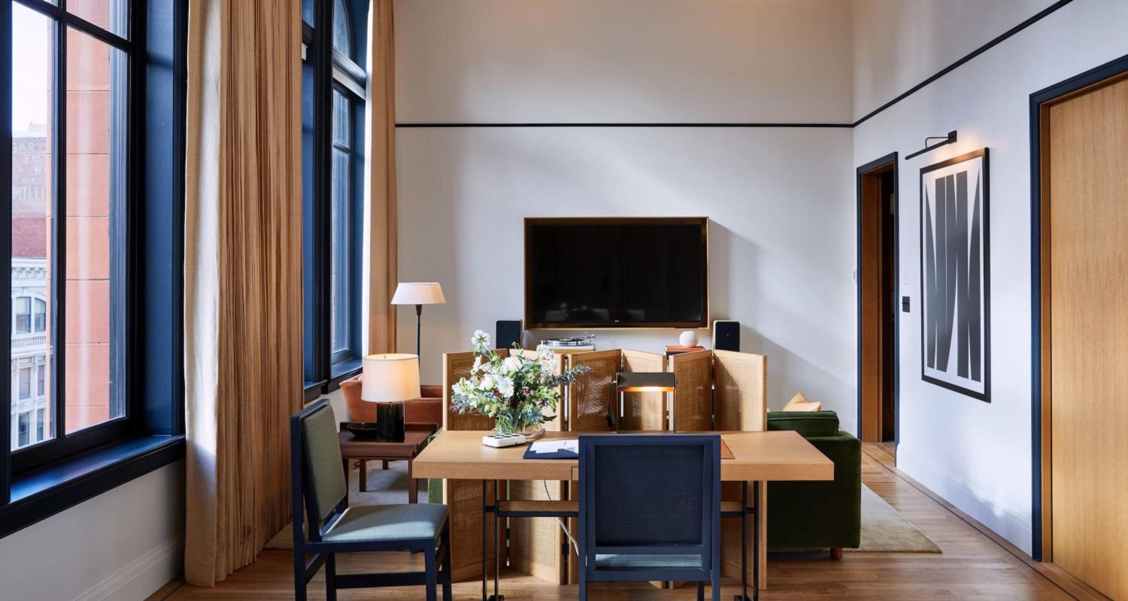 A hotel room with a wooden dining table, two chairs, a sofa, a TV mounted on the wall, and a window with curtains.