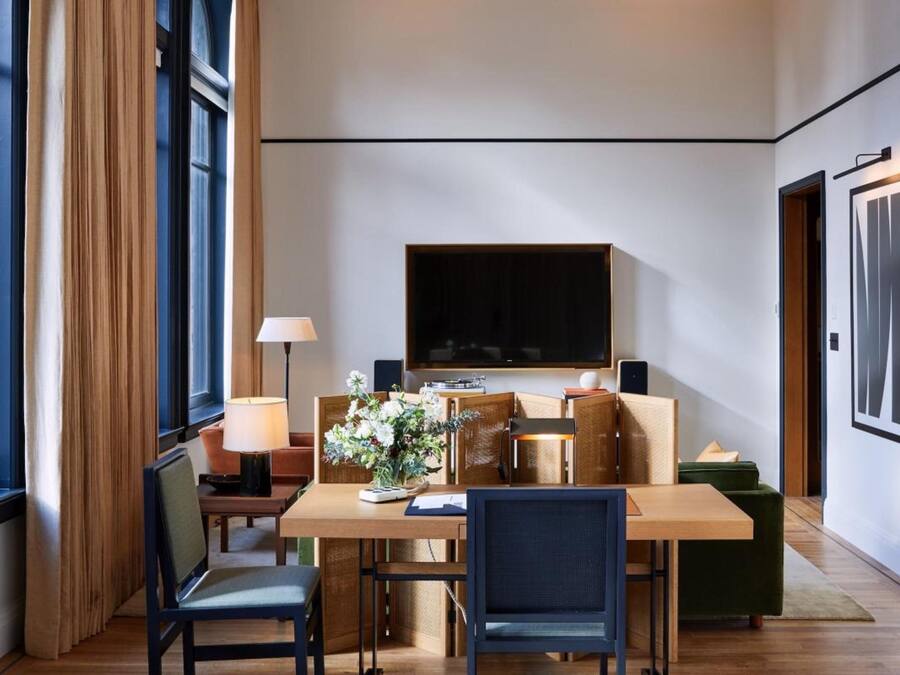 A hotel room with a wooden dining table, two chairs, a sofa, a TV mounted on the wall, and a window with curtains.