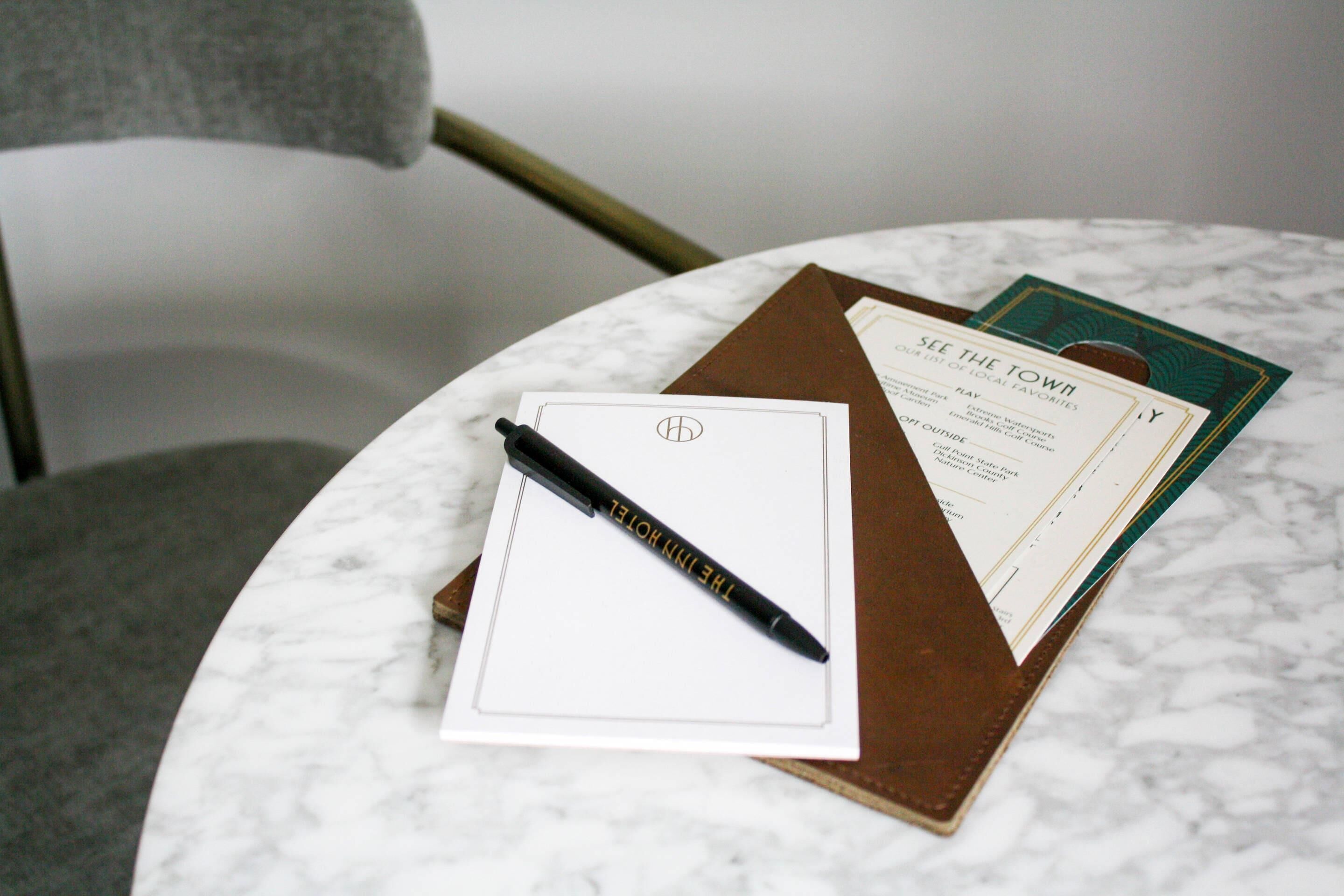 Un cuaderno con un bolígrafo sobre una mesa de mármol, una carpeta marrón y un folleto con el título 'SEE THE TOWN'.