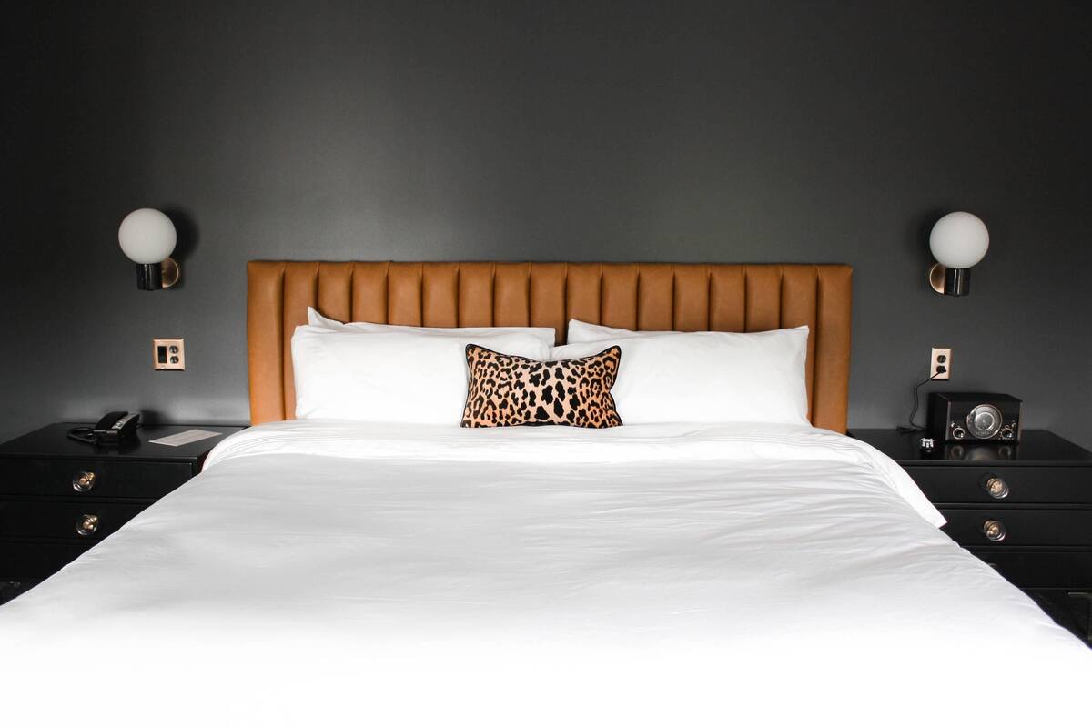 Una cama bien hecha con sábanas blancas y una única almohada de estampado de leopardo, flanqueada por mesitas de noche negras con una lámpara de mesa redonda y otra lámpara pequeña.