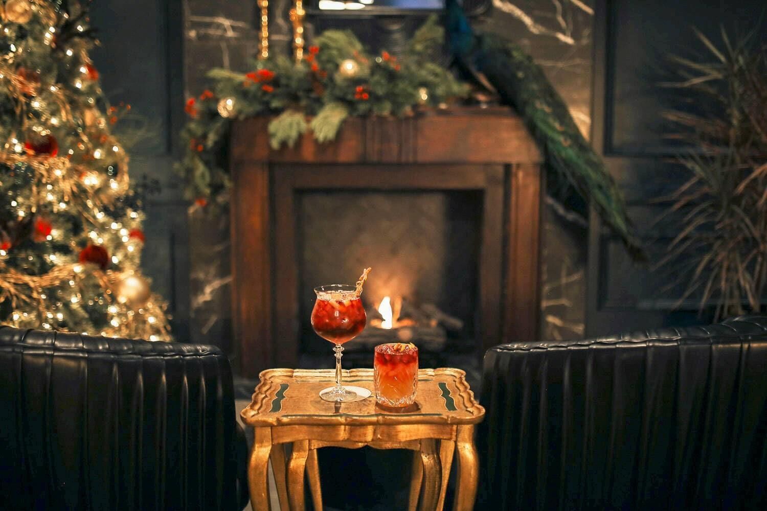 A cozy seating area with a small table featuring two drinks, a decorated Christmas tree, and a fireplace in the background.
