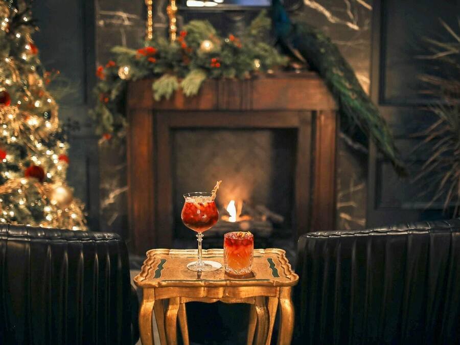 A cozy seating area with a small table featuring two drinks, a decorated Christmas tree, and a fireplace in the background.