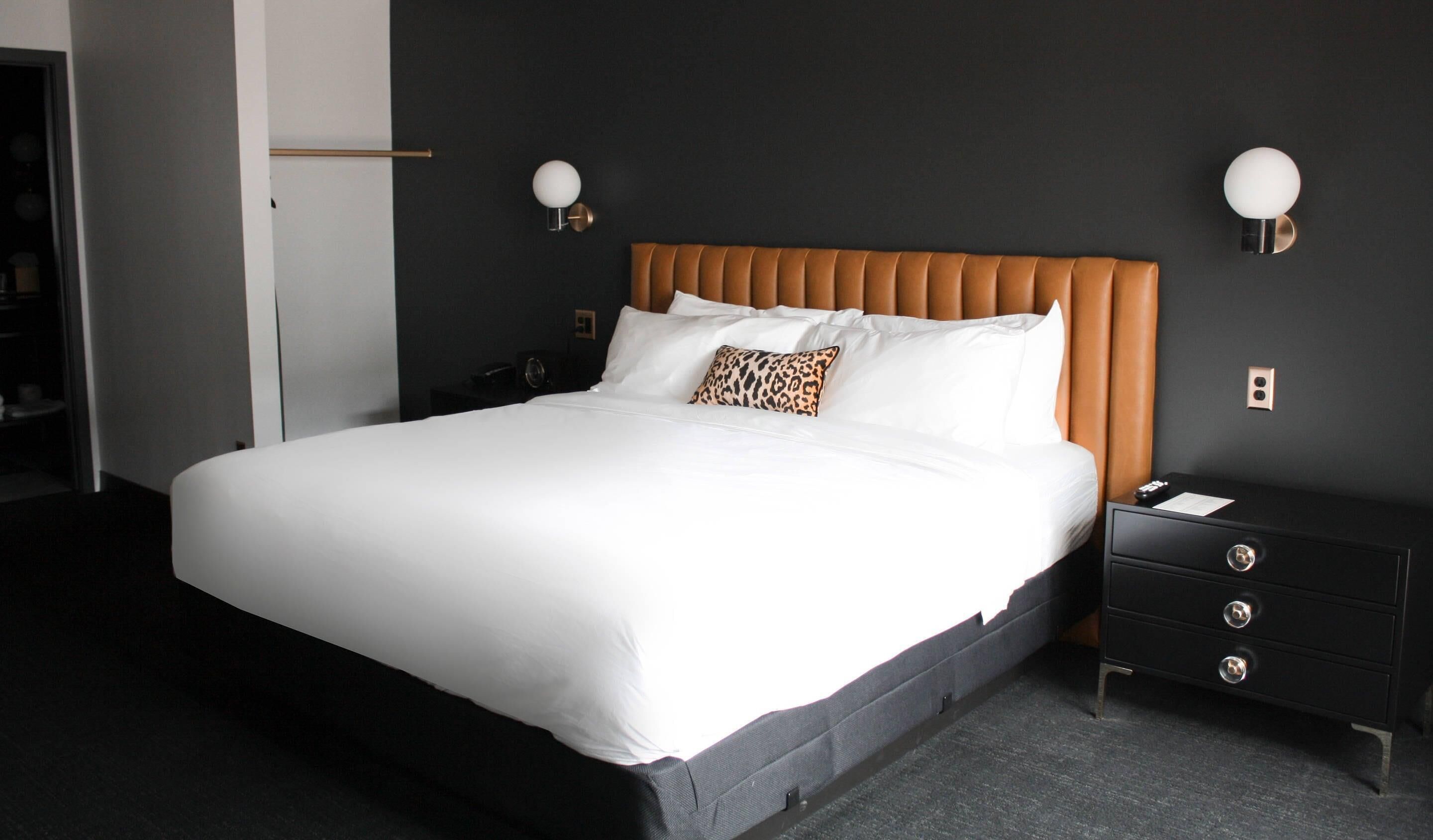 A neatly made bed with white linens and a single leopard print pillow, a wooden headboard, and a black nightstand with drawers.