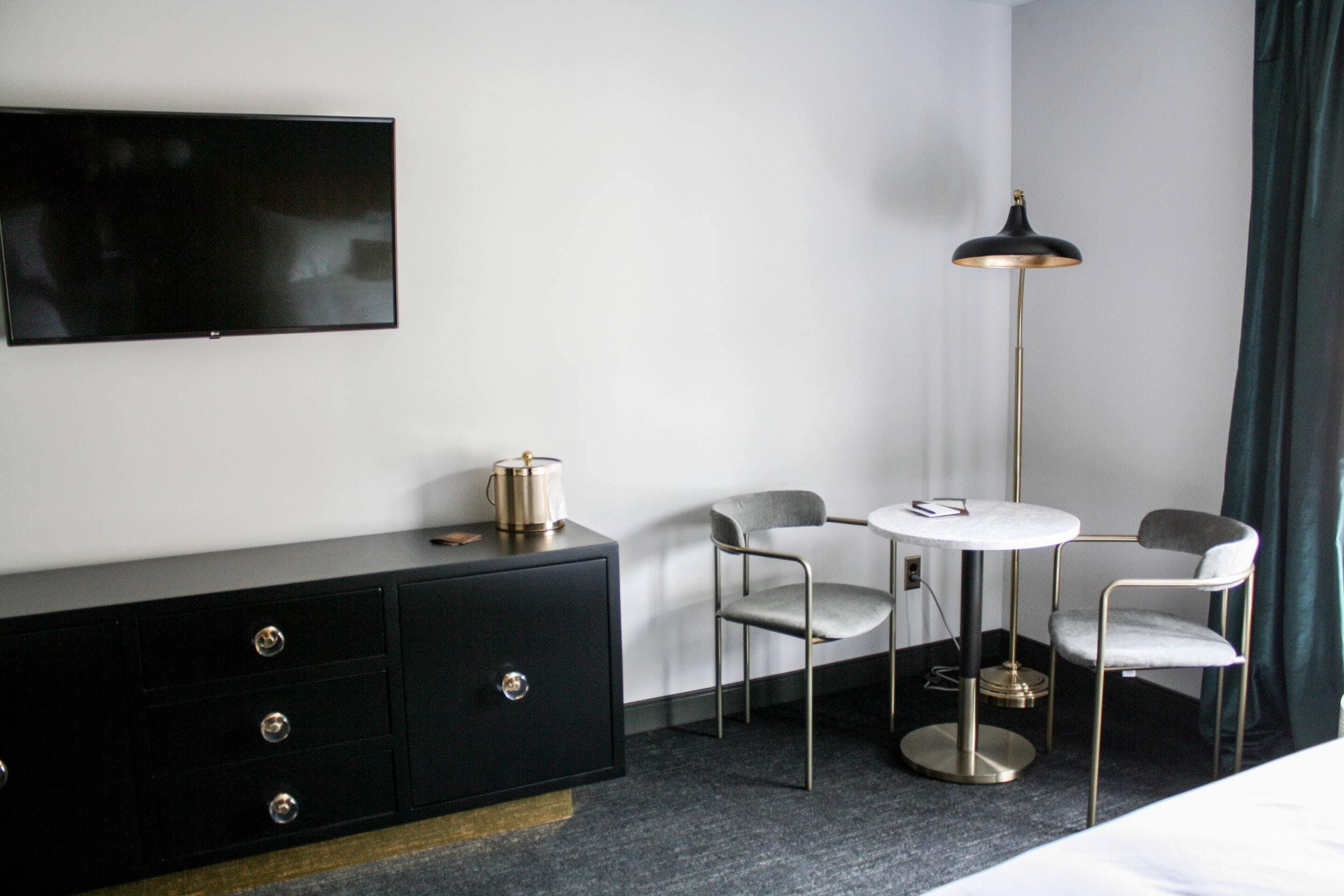A hotel room with a flat-screen TV, a black dresser, two chairs, a small round table, and a floor lamp.
