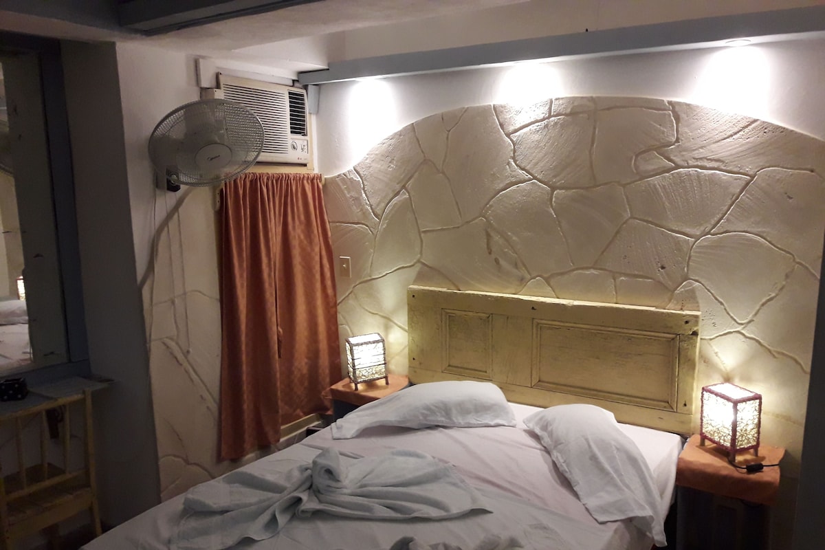 A bedroom with a stone headboard, a bed with white linens, a standing fan, a heater, and a nightstand with a lamp.