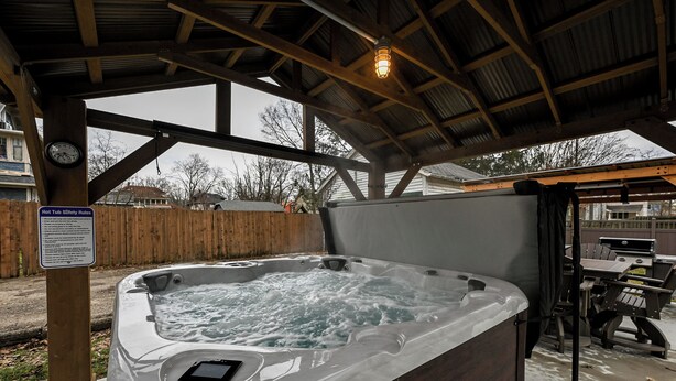 Un jacuzzi bajo una estructura de madera, con un cartel de seguridad, un reloj y una parrilla al fondo.
