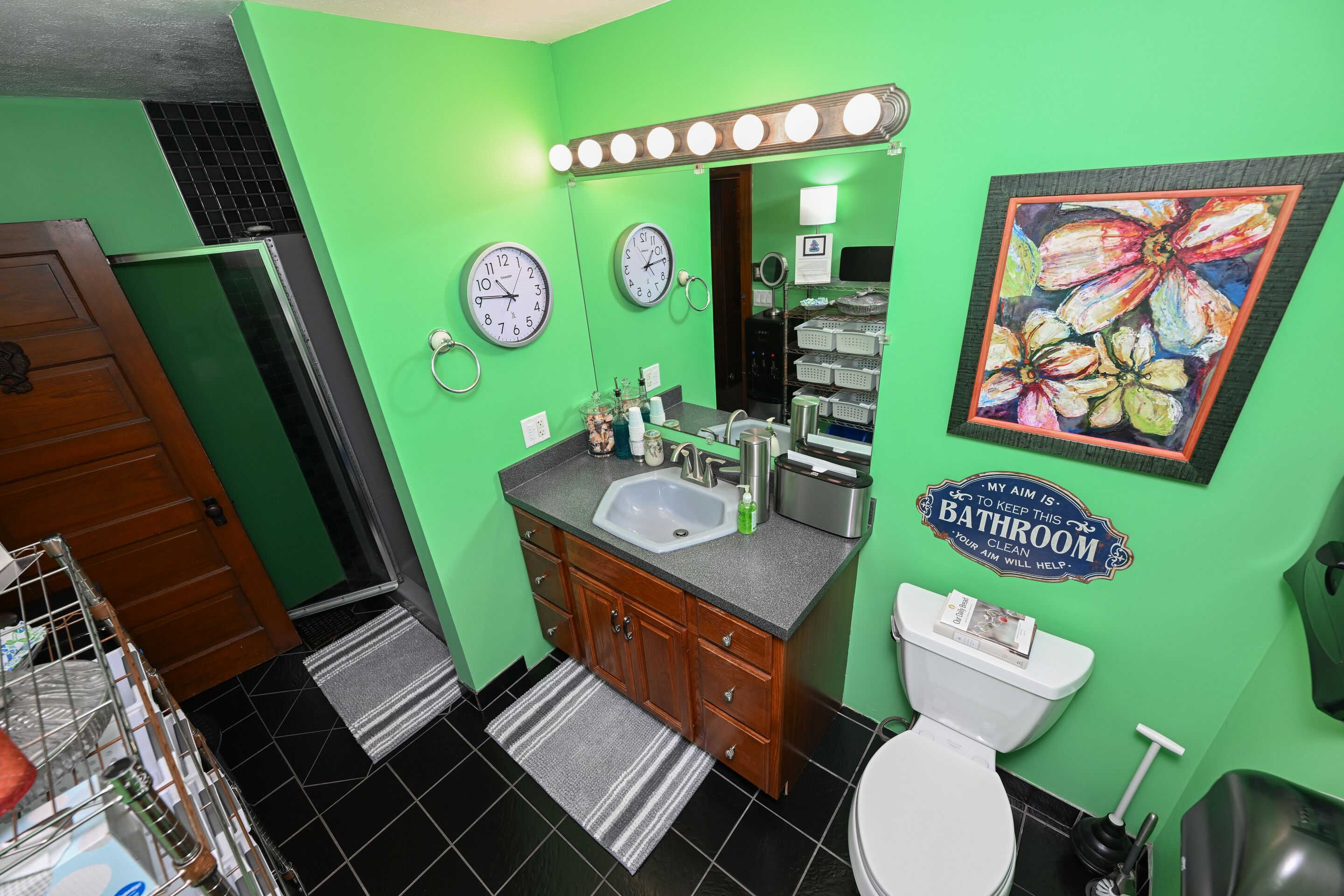 Une salle de bain avec des murs verts, un lavabo, des toilettes et un miroir avec des lumières au-dessus. Il y a des horloges au mur et une pancarte qui indique : « Mon objectif est de maintenir cette salle de bain propre. »
