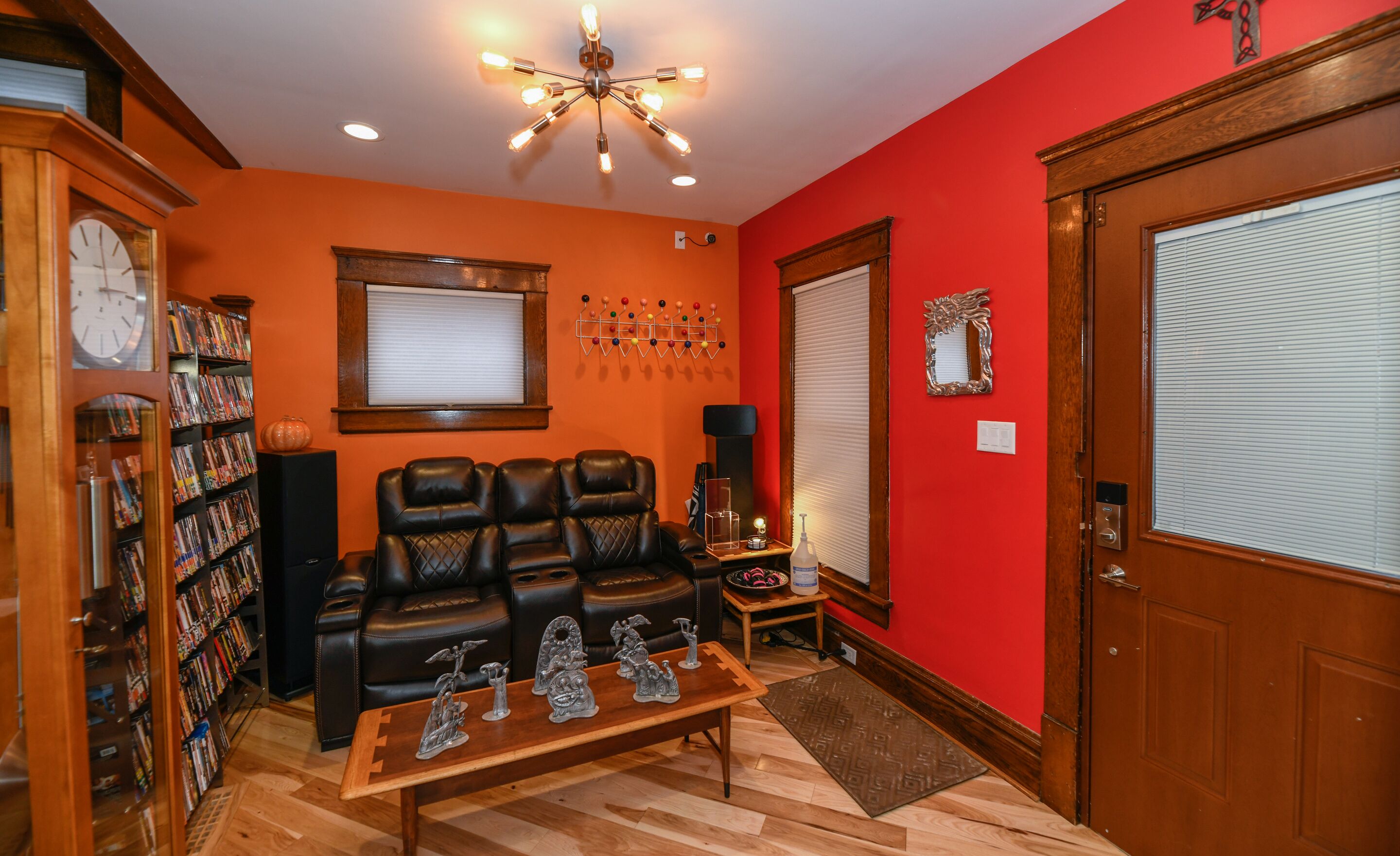 A living room with a red wall, a wooden door, a bookshelf, a leather sofa, a coffee table with figurines, and a ceiling light fixture.