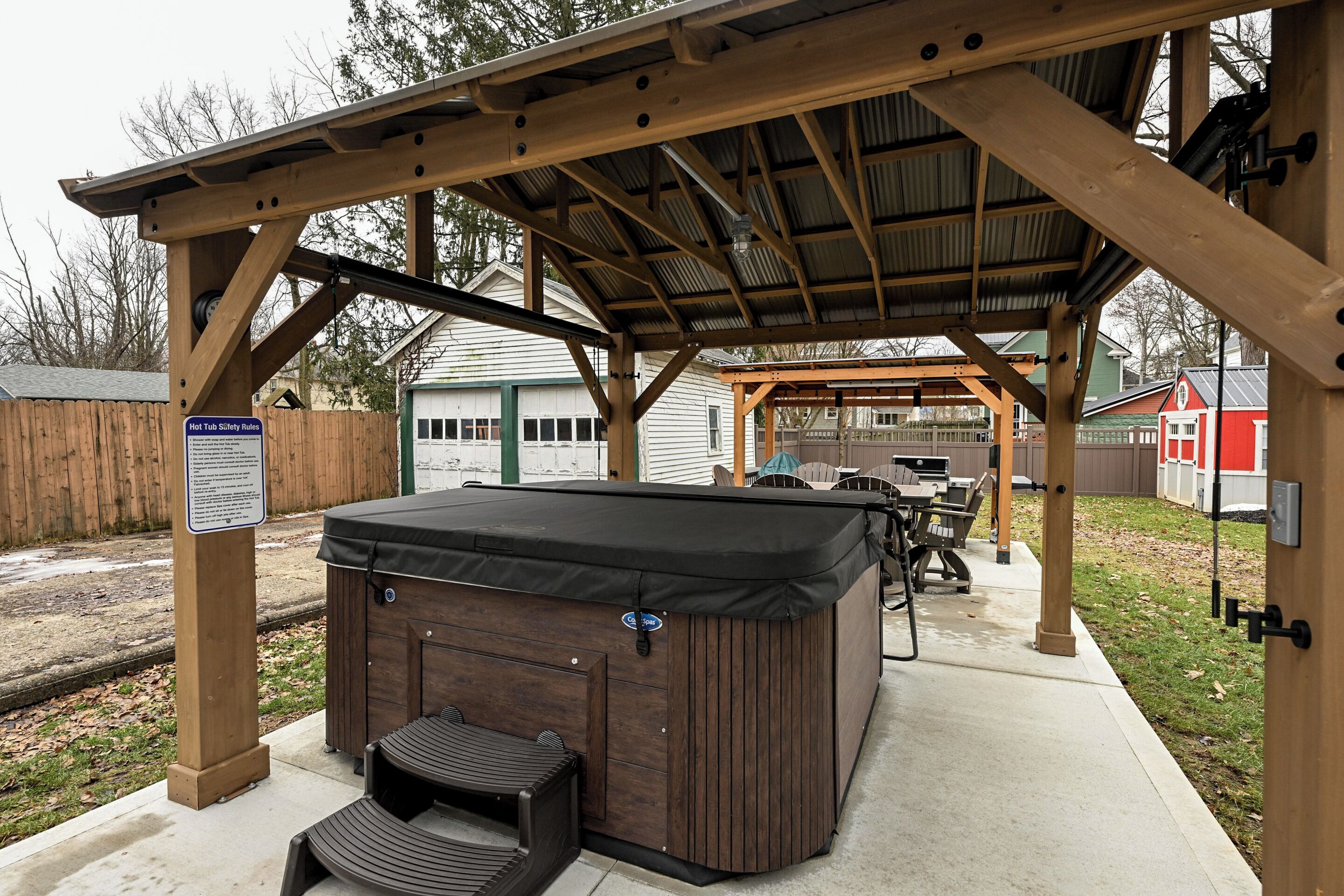 A hot tub under a wooden pergola with a covered patio area, surrounded by a fenced yard and a red building in the background.