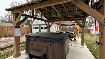 A hot tub under a wooden pergola with a covered patio area, surrounded by a fenced yard and a red building in the background.