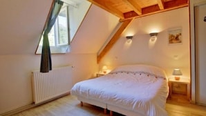 Ein Schlafzimmer mit schrägen Wänden, einem Bett, zwei Nachttischlampen, einem Heizkörper und einem Fenster mit Vorhängen.