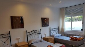 Ein Hotelzimmer mit drei Einzelbetten, einem hölzernen Nachttisch, einem kleinen Tisch und einem Fenster mit Jalousien.